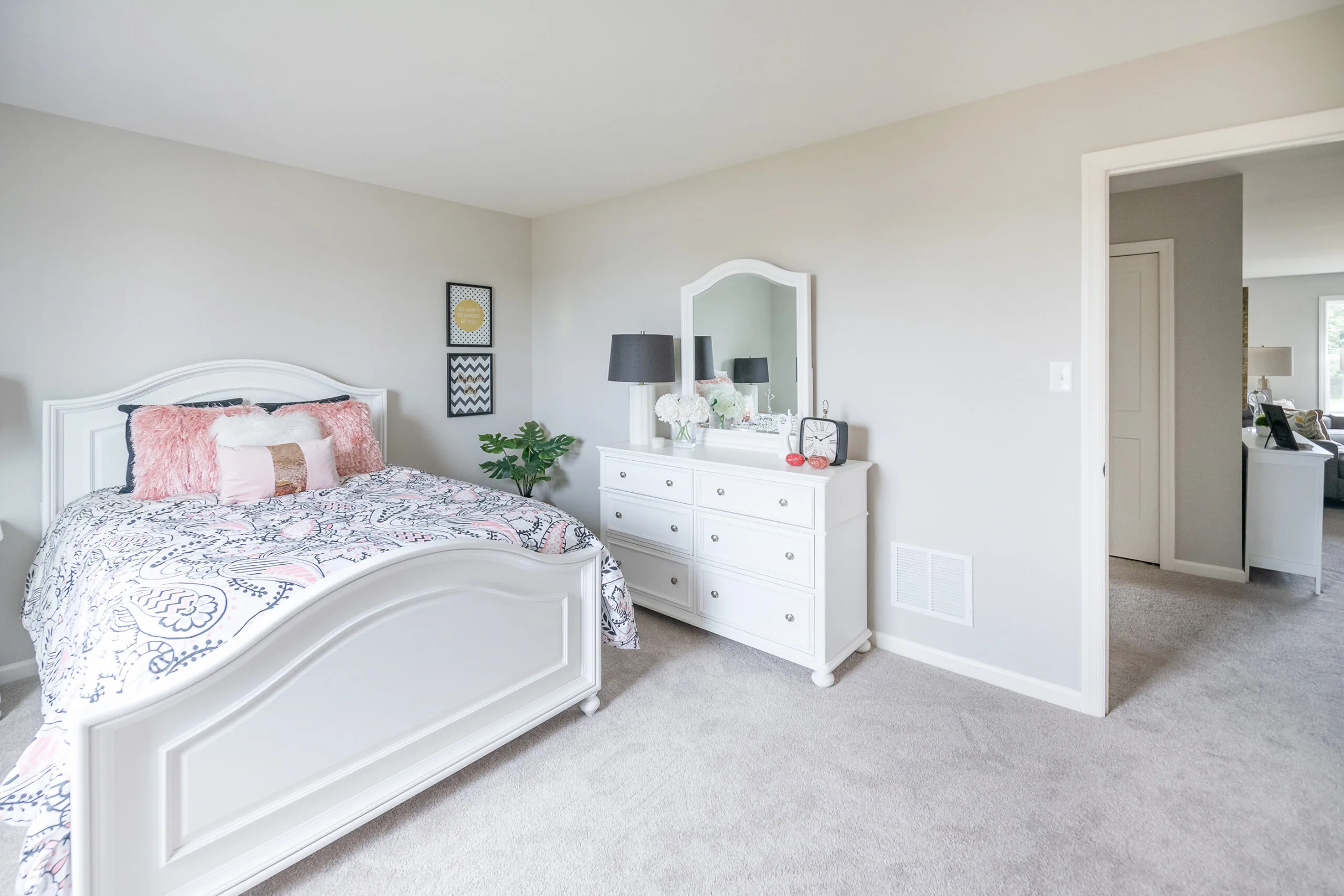 A bright bedroom with light-colored walls, a white bed with patterned bedding and pink pillows, a matching white dresser with a mirror, two black lamps, and decorative items. A doorway leads to another room.