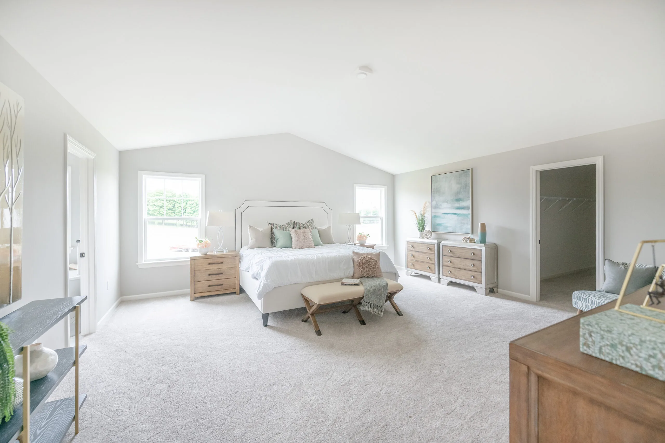 A bright, spacious bedroom with a white bed, two nightstands, a bench at the foot of the bed, dressers, wall art, large windows, and light carpeting. The decor is modern and minimalistic in neutral tones.