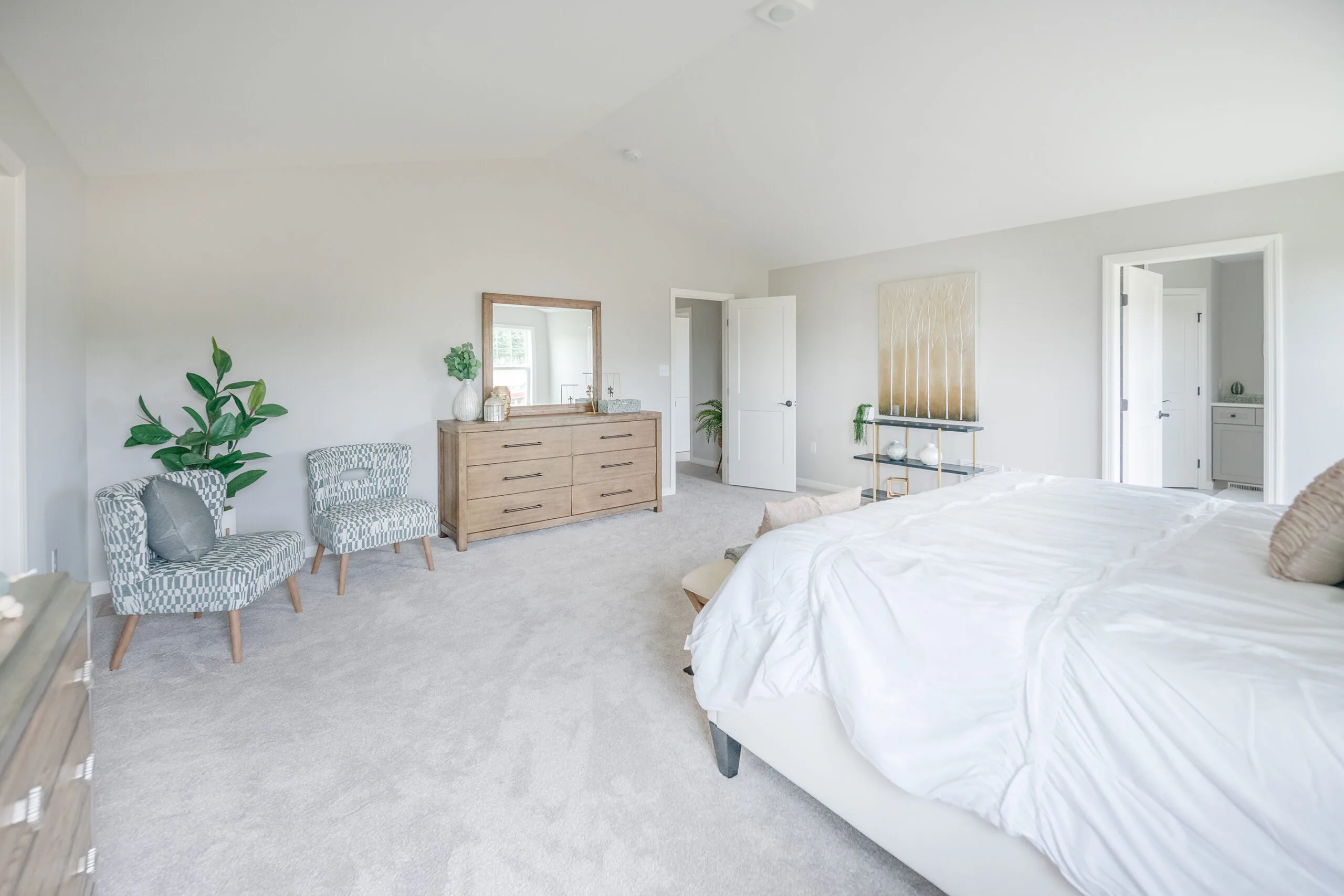 Bright, spacious bedroom with light gray carpet, a large bed with white bedding, two patterned chairs, a wooden dresser with a mirror, potted plants, and white walls with minimal decor.