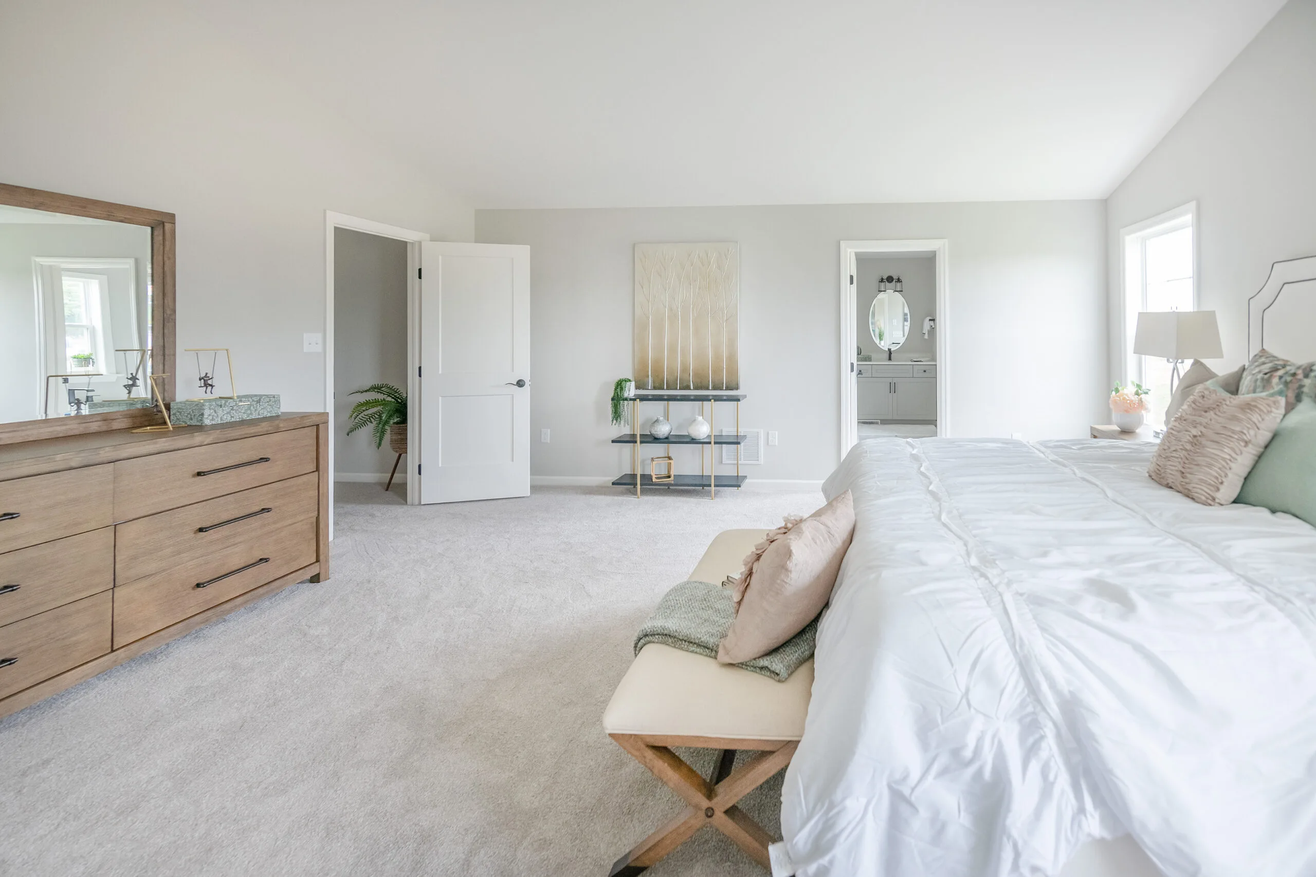 Spacious, bright bedroom with a large bed, bench, wooden dresser, and soft gray carpet. Open doors lead to a bathroom and another room. Wall art, mirrors, and windows enhance the airy, modern decor.