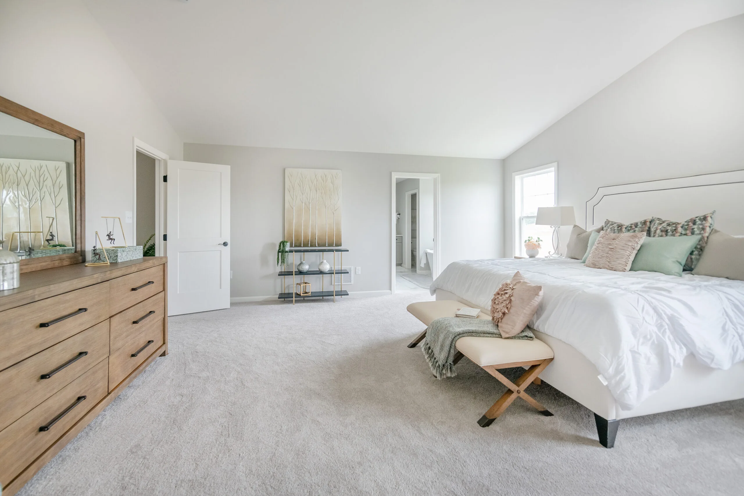 Spacious, bright bedroom with a neatly made bed, decorative pillows, a bench at the foot, a wooden dresser with a mirror, soft carpet, and light-colored walls. Sunlight streams in through a window, creating a cozy atmosphere.