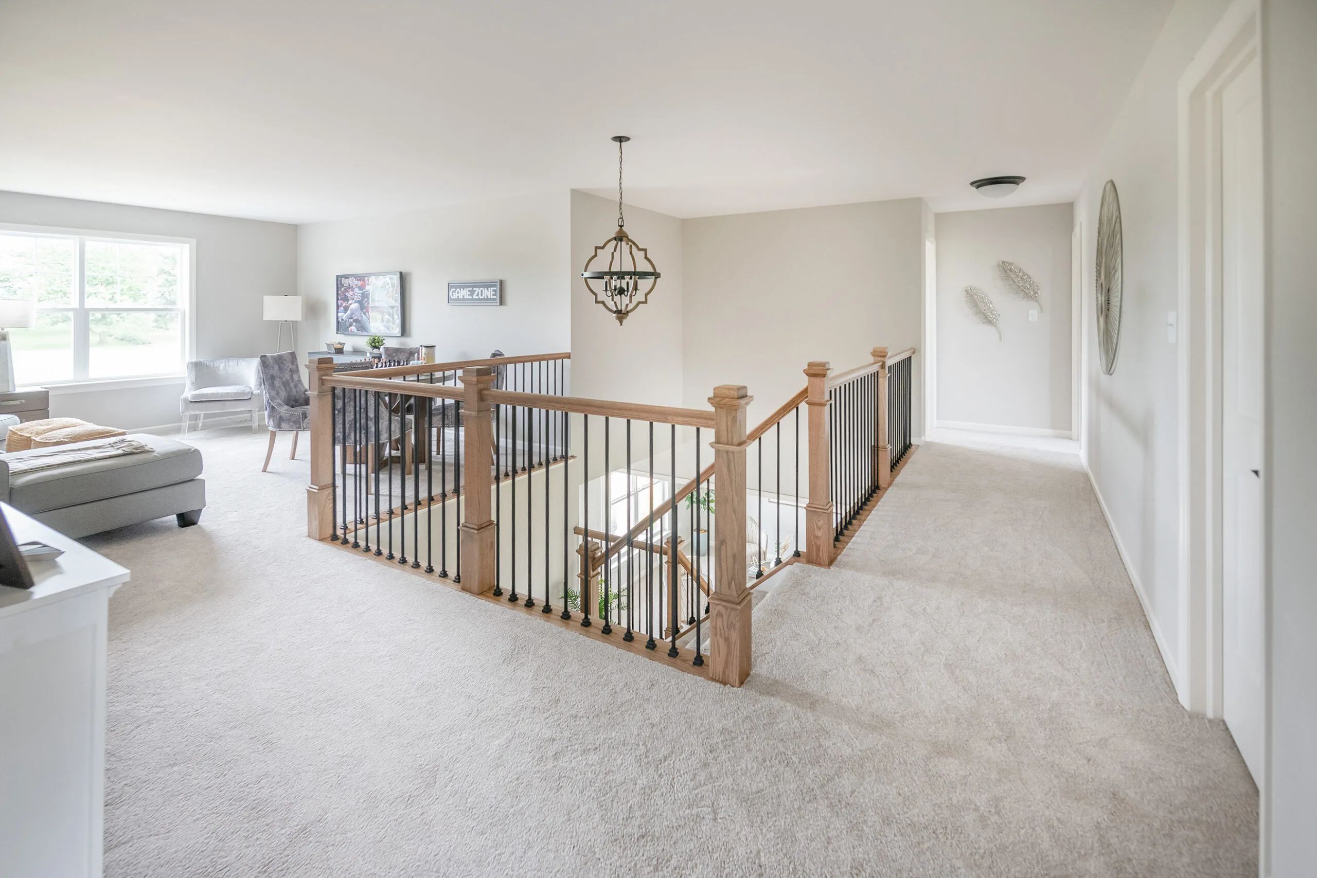 A spacious, bright upstairs hallway with light carpet, wooden railing with black spindles, a modern chandelier, and a sitting area by a large window with a sofa, chair, and desk. White walls and decor create an airy feel.