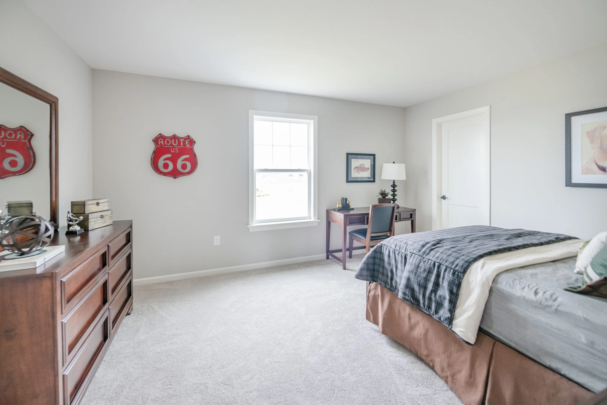 A bright bedroom with light walls, a neatly made bed, a wooden dresser, a desk with a chair by the window, and a red Route 66 sign hanging on the wall. The room looks tidy and spacious.