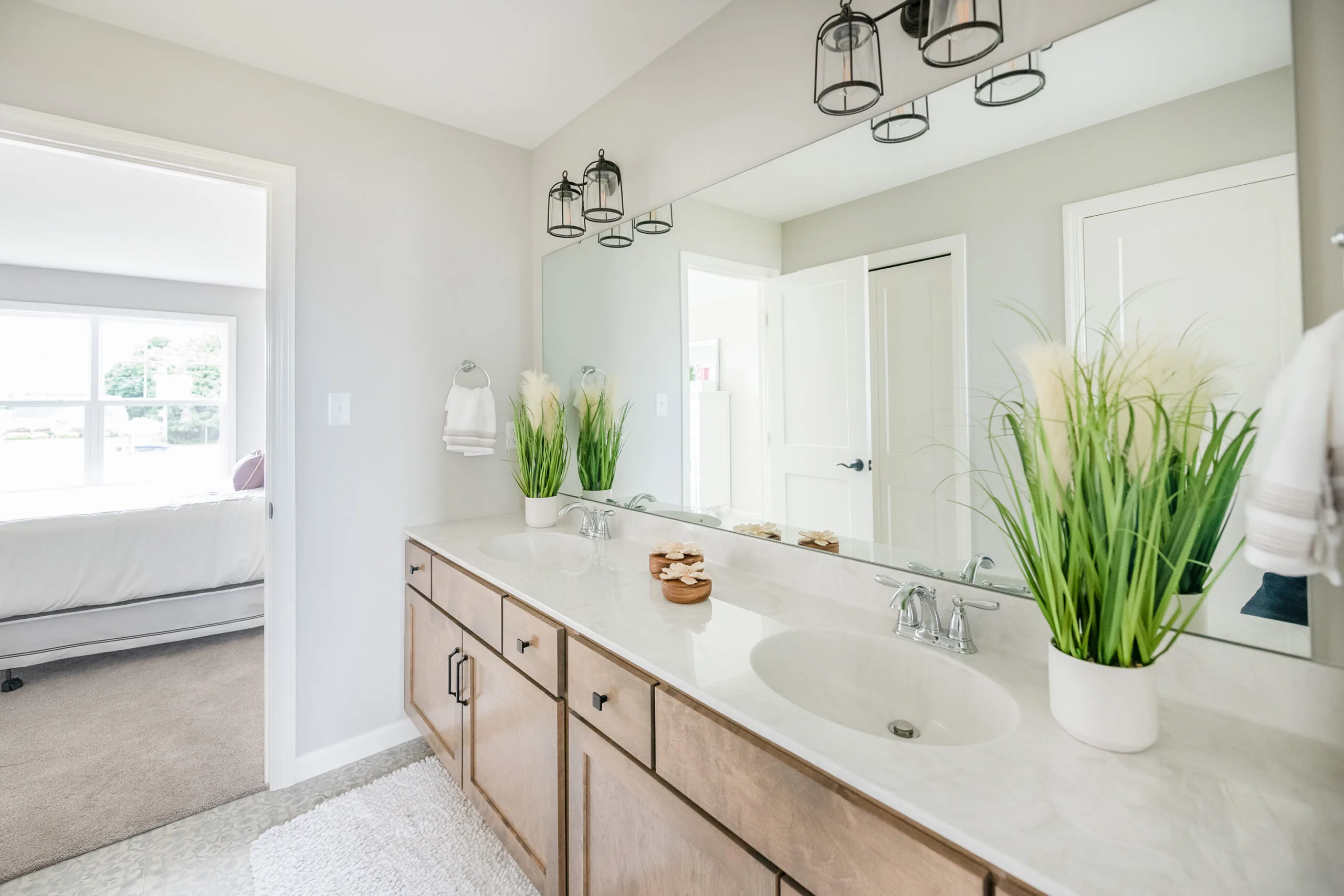 Bright, modern bathroom with a large mirror, double sinks, wooden cabinets, and two tall plants on the counter. White walls, towel racks, and a glimpse of a bedroom through an open door.