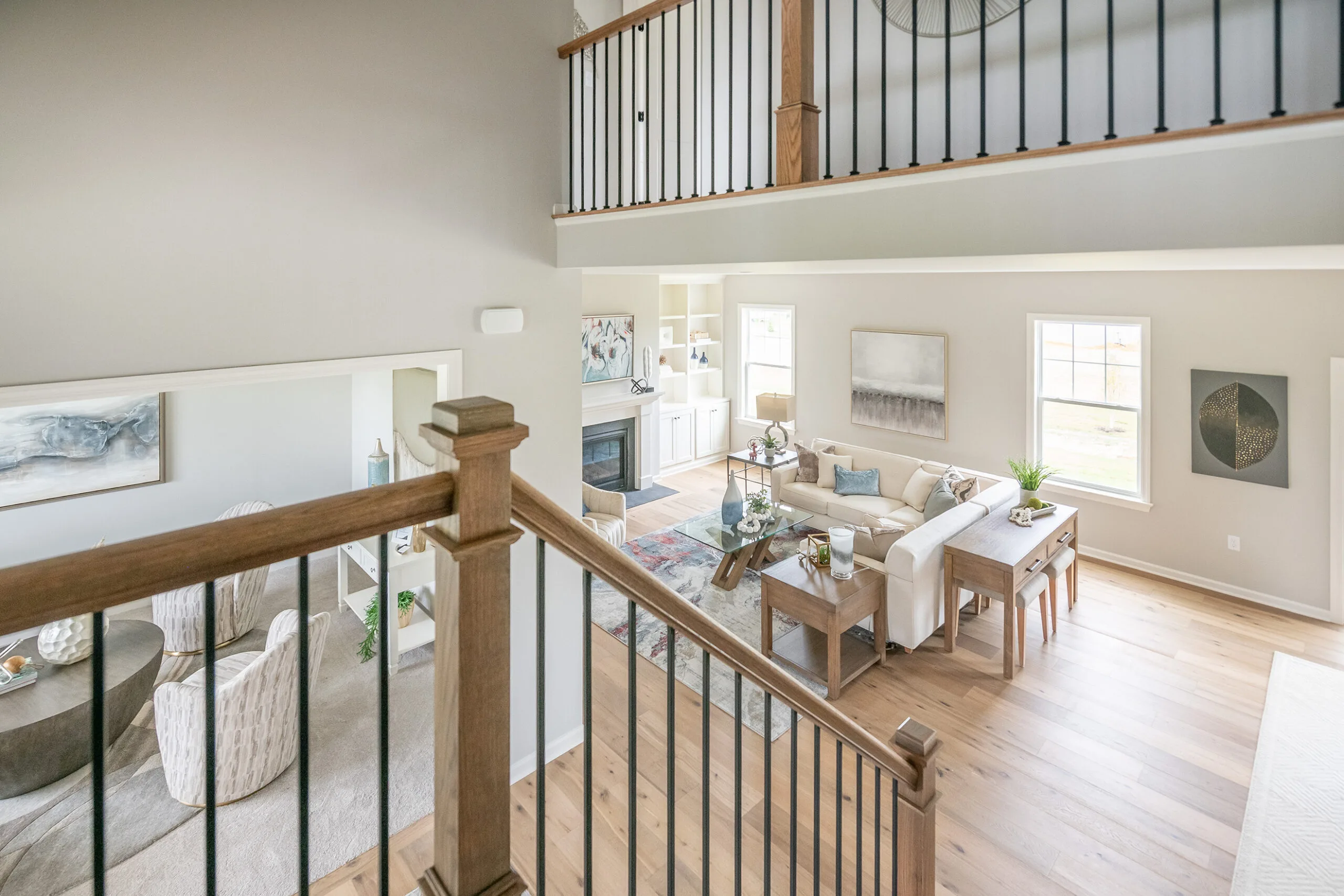 A view from an upper staircase landing overlooks a bright, open living room with light wood flooring, neutral furniture, large windows, and adjacent sitting area. Natural light fills the modern and inviting space.