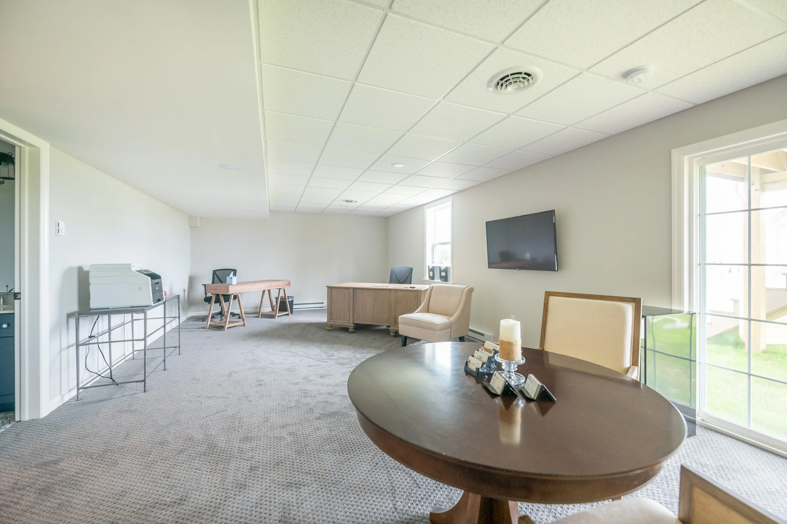 A spacious home office with neutral walls, carpeted floor, desks, chairs, a round table with decor, a wall-mounted TV, and a large window letting in natural light.