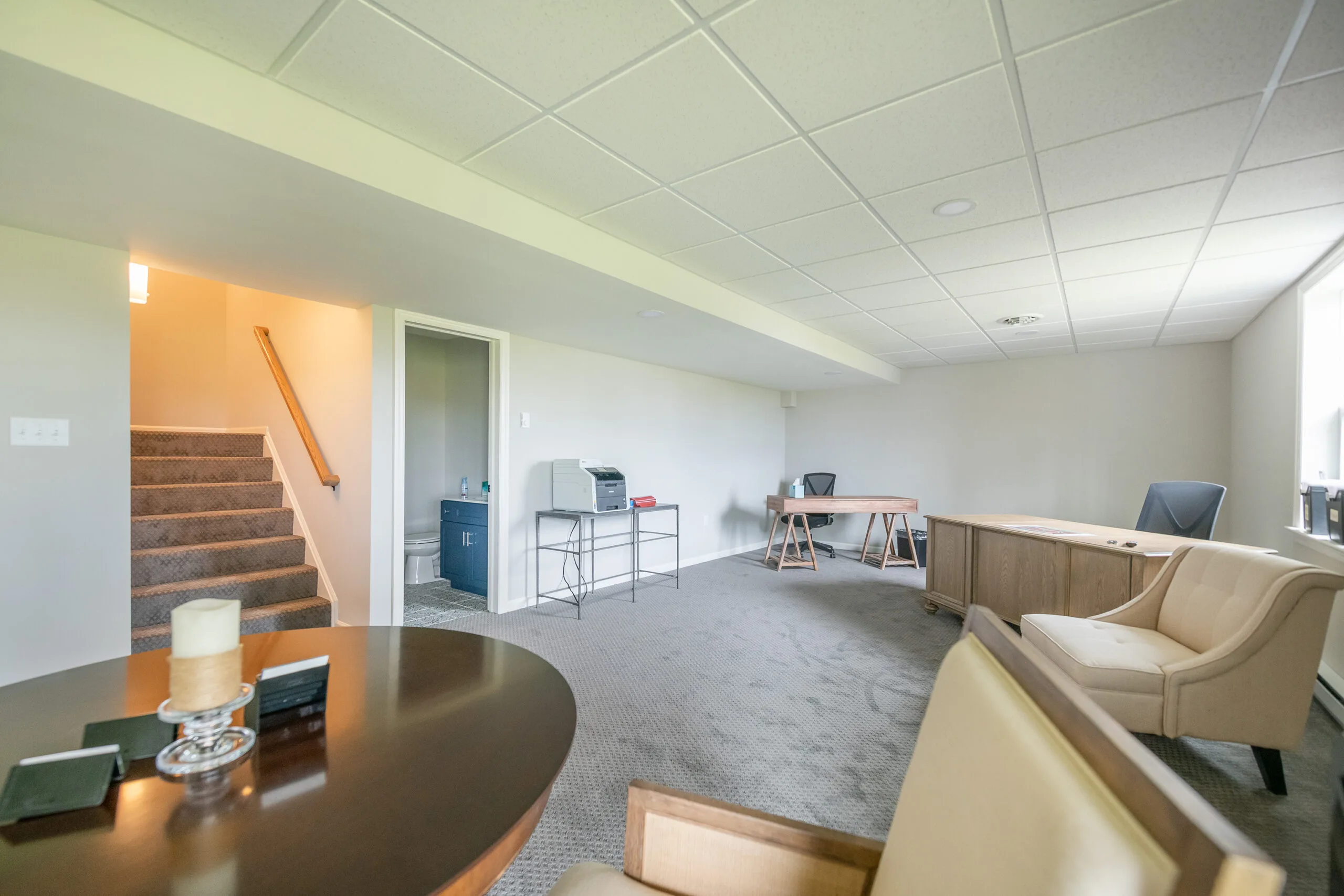 A spacious, carpeted basement office with desks, chairs, and office equipment. Stairs lead up on the left, and a small table with a candle is in the foreground. Light streams in from windows on the right.