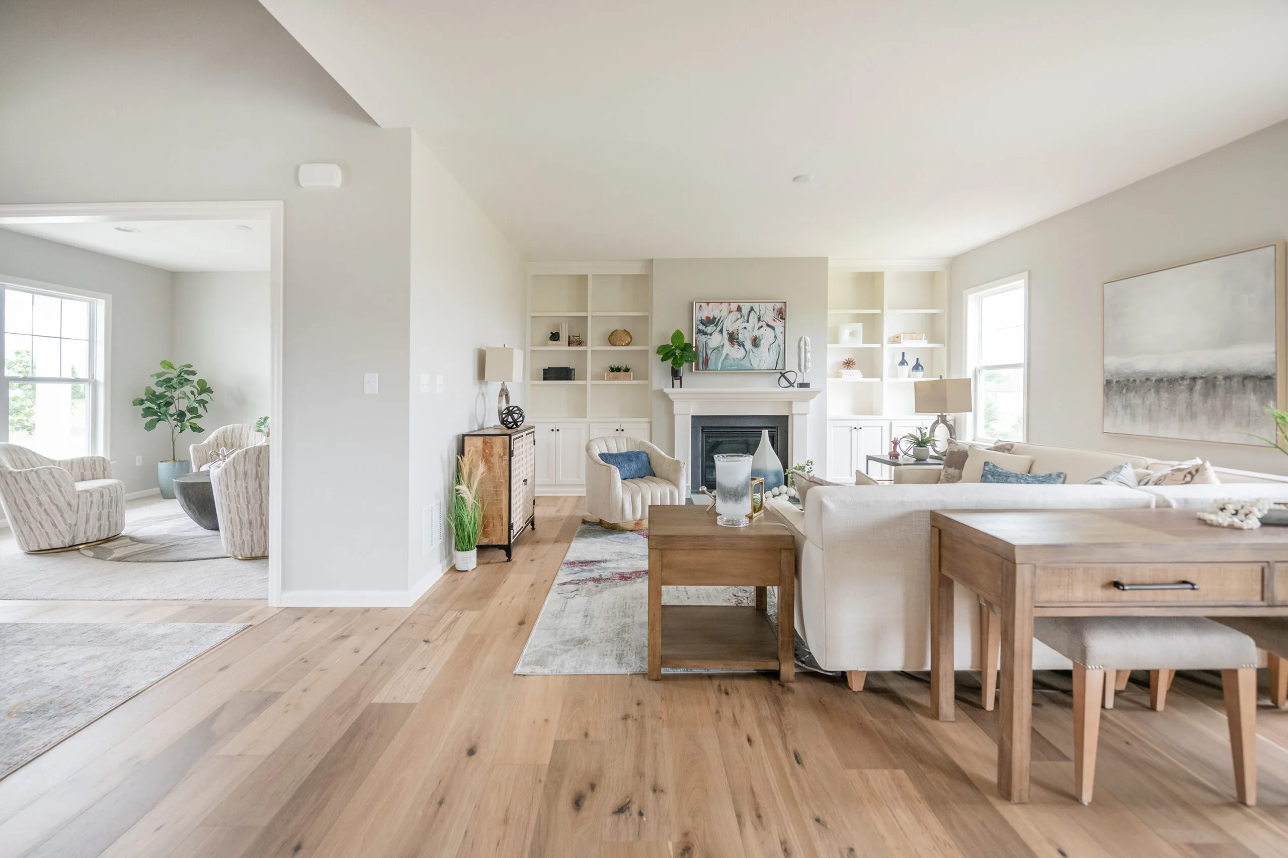 Bright, modern living room with light wood floors, white walls, built-in shelves, a fireplace with art above, and neutral furniture. An adjacent sitting area is visible through an open doorway. Natural light fills the space.