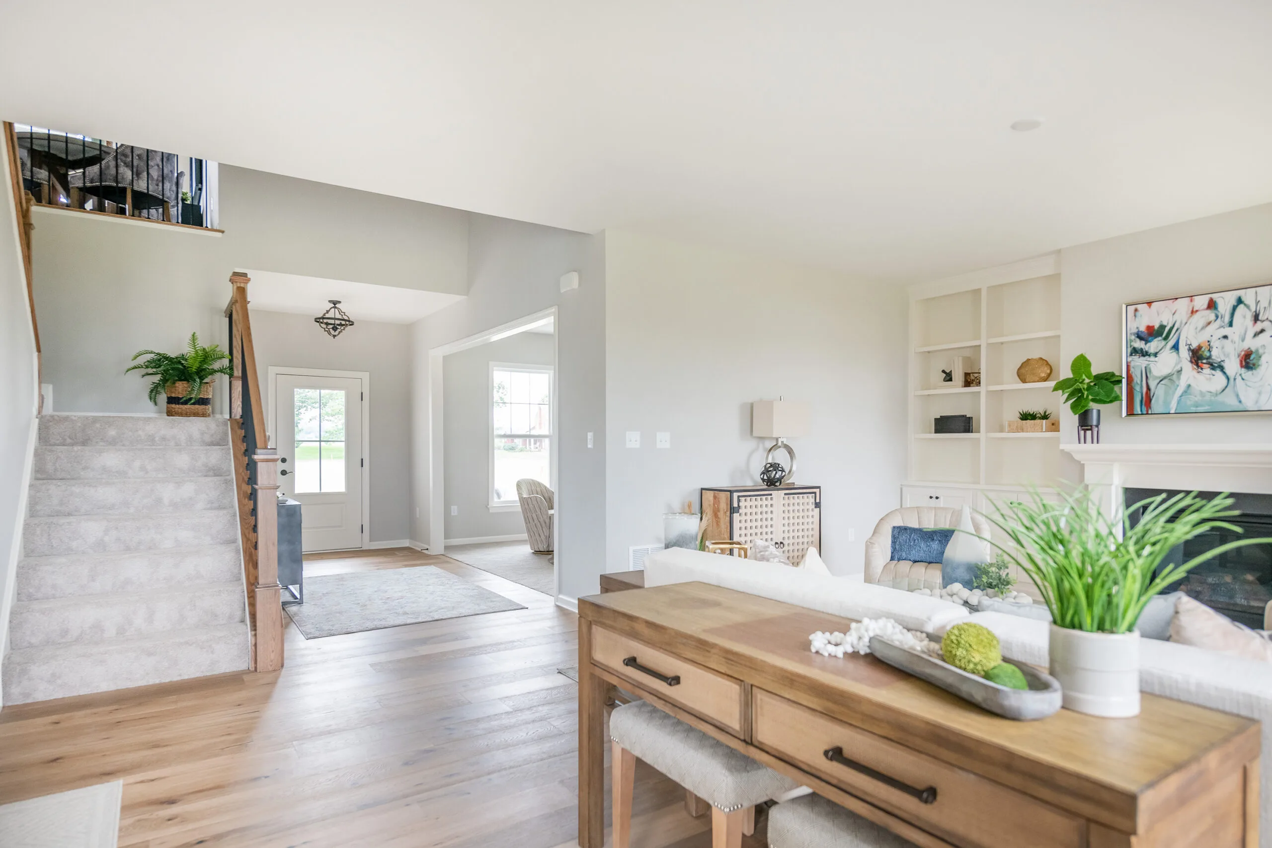 Bright, modern living room with light wood floors, white walls, built-in shelves, and a cozy seating area. Stairs with a carpet runner lead up, and a console table with plants sits behind the sofa. Natural light fills the space.