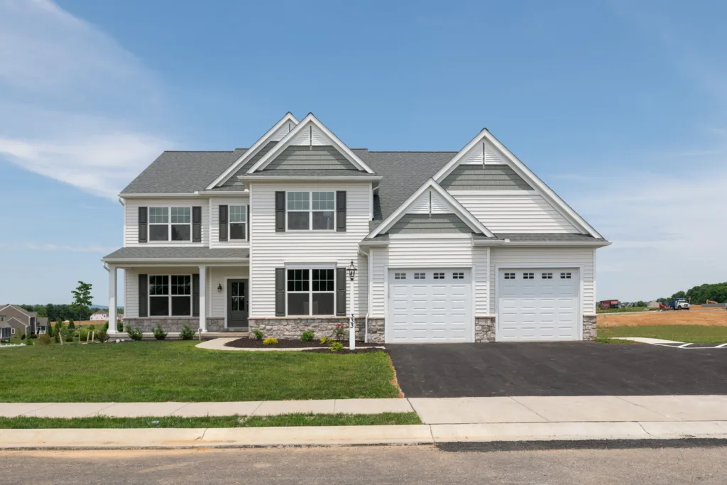 A modern two-story suburban house with white siding, gray roof shingles, a front porch, and a double garage, situated on a well-kept lawn with a paved driveway on a clear, sunny day.