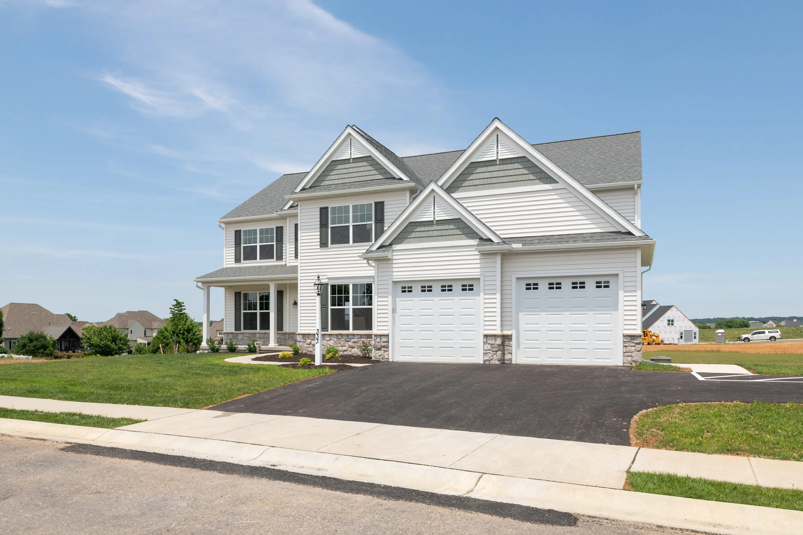 A modern two-story suburban house with white siding, a double garage, large windows, and a neatly manicured lawn under a clear blue sky. Other houses and open fields are visible in the background.