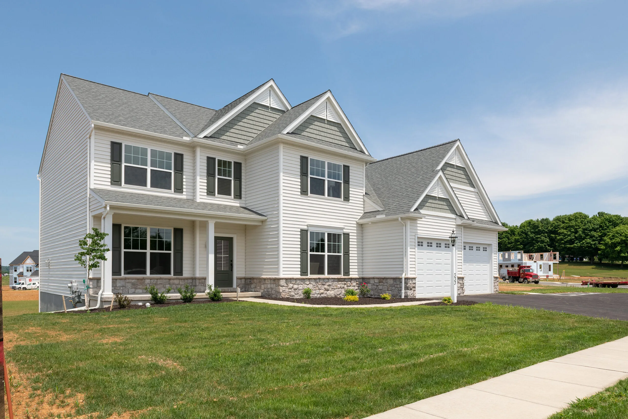 A two-story suburban house with white siding, gray roof, double garage, and large front lawn on a sunny day. There are small shrubs along the house and a driveway leading to the garage.
