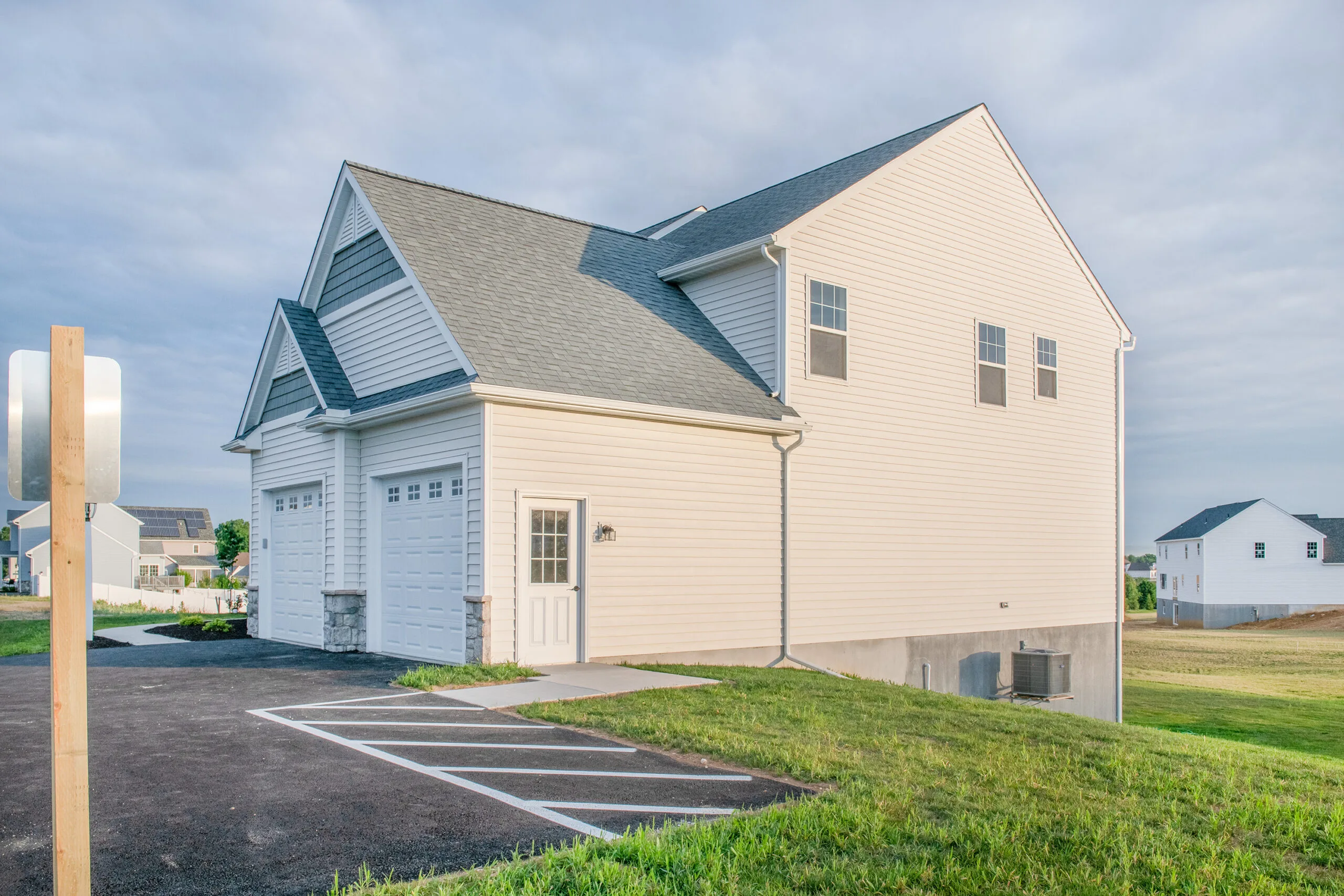 A beige, two-story suburban house with a double garage and a white door, adjacent to a newly paved driveway with marked parking spaces, surrounded by green grass and neighboring houses in the background.