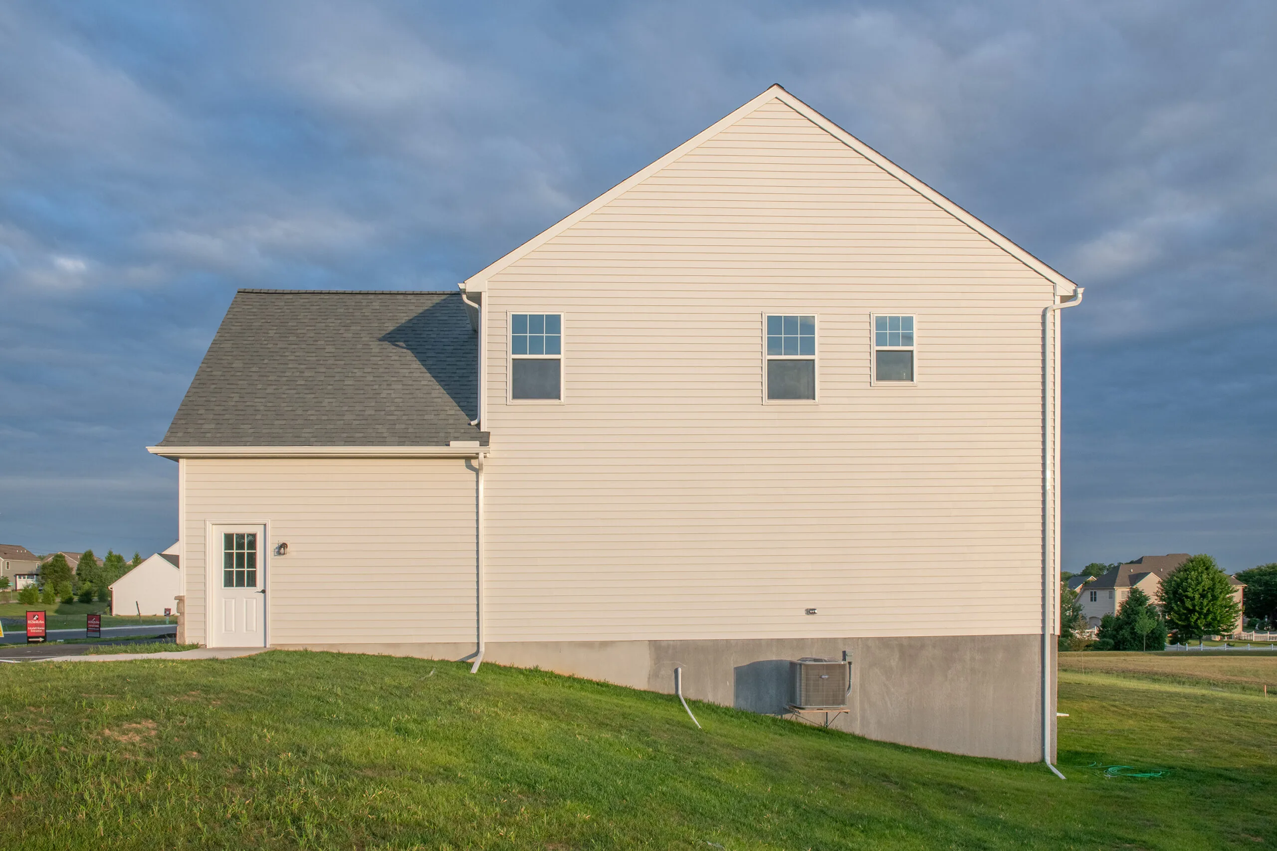 A beige house with three windows and a side door sits on a grassy slope under a cloudy sky, with suburban homes visible in the background.