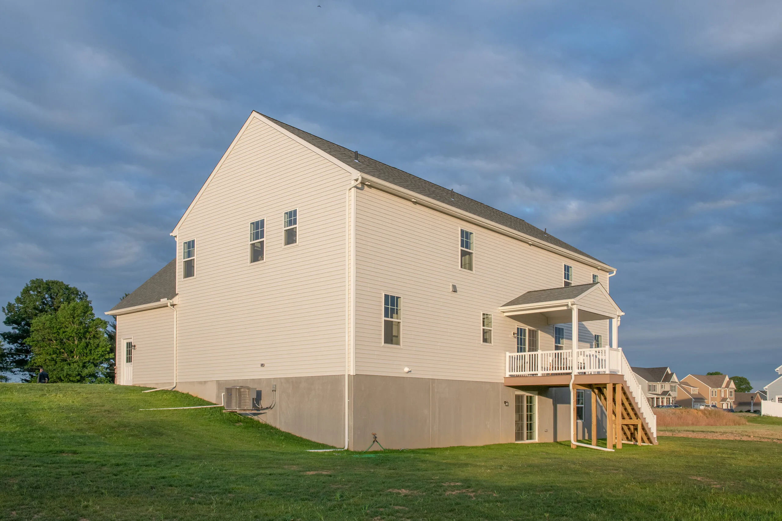 A large, two-story beige house with a gray foundation, elevated deck with stairs, multiple windows, and a sloped grassy lawn under a cloudy sky.