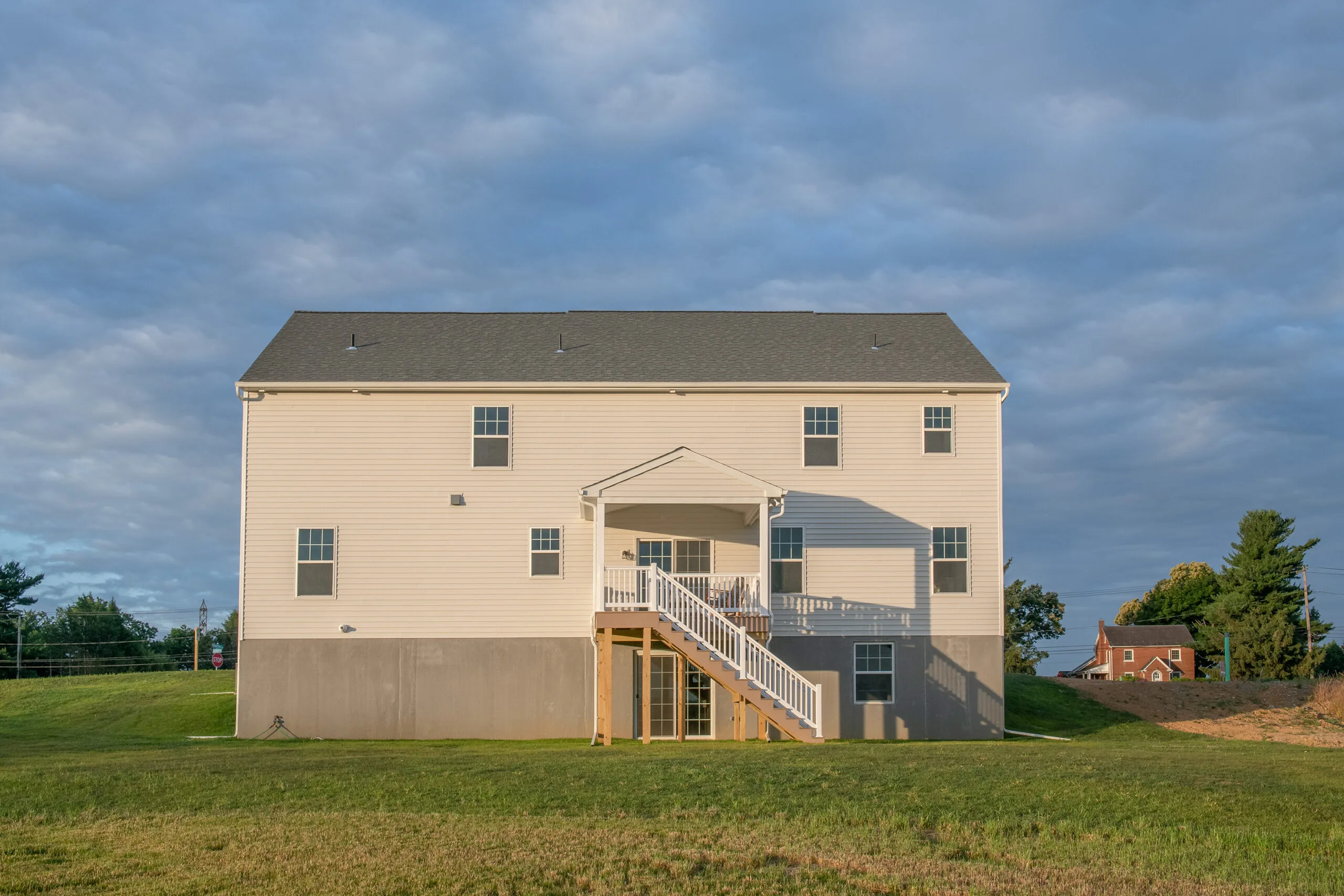 A large, beige two-story house with a covered porch and exterior staircase leading to the backyard, set on a grassy lawn under a partly cloudy sky.