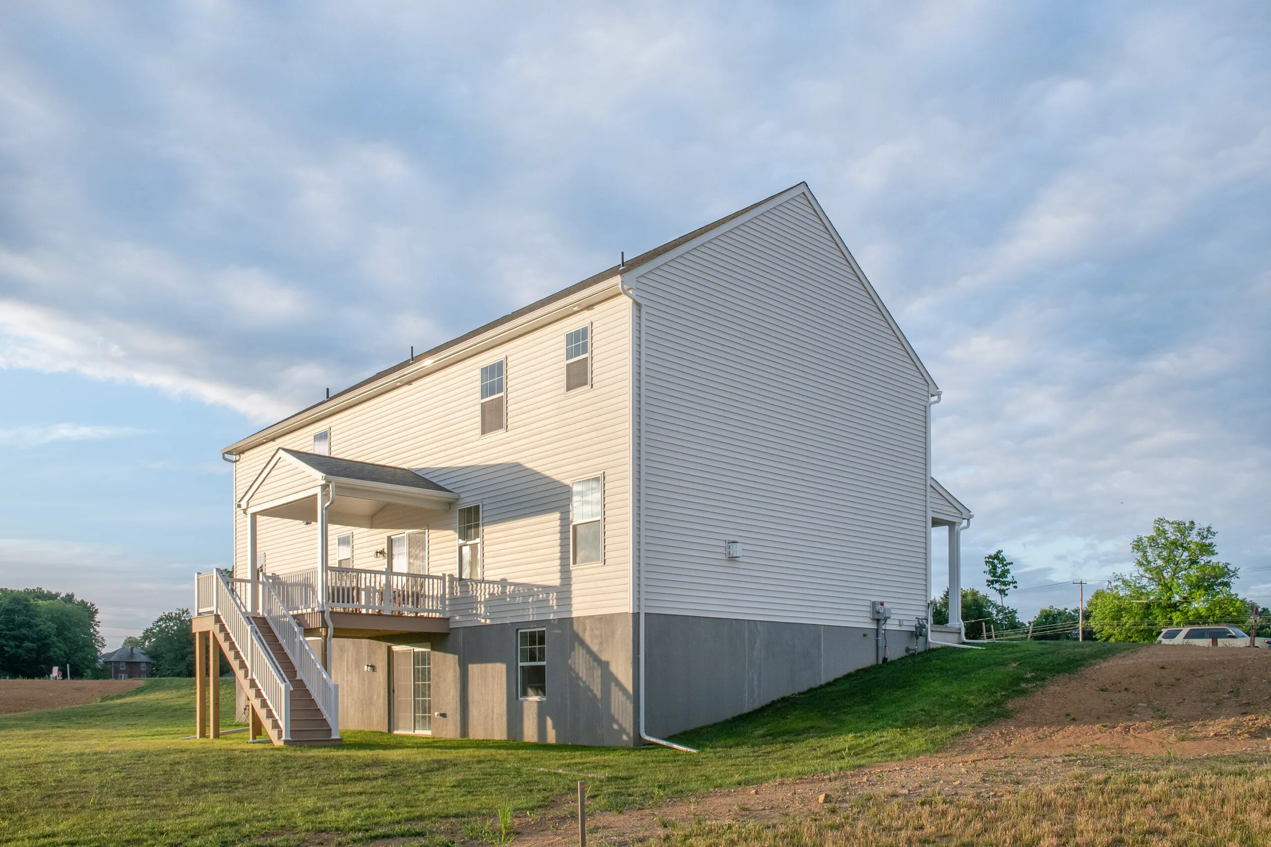 A two-story white house with a covered back porch and stairs leading to a grassy backyard, set against a partly cloudy sky. The house has large windows and a concrete foundation.