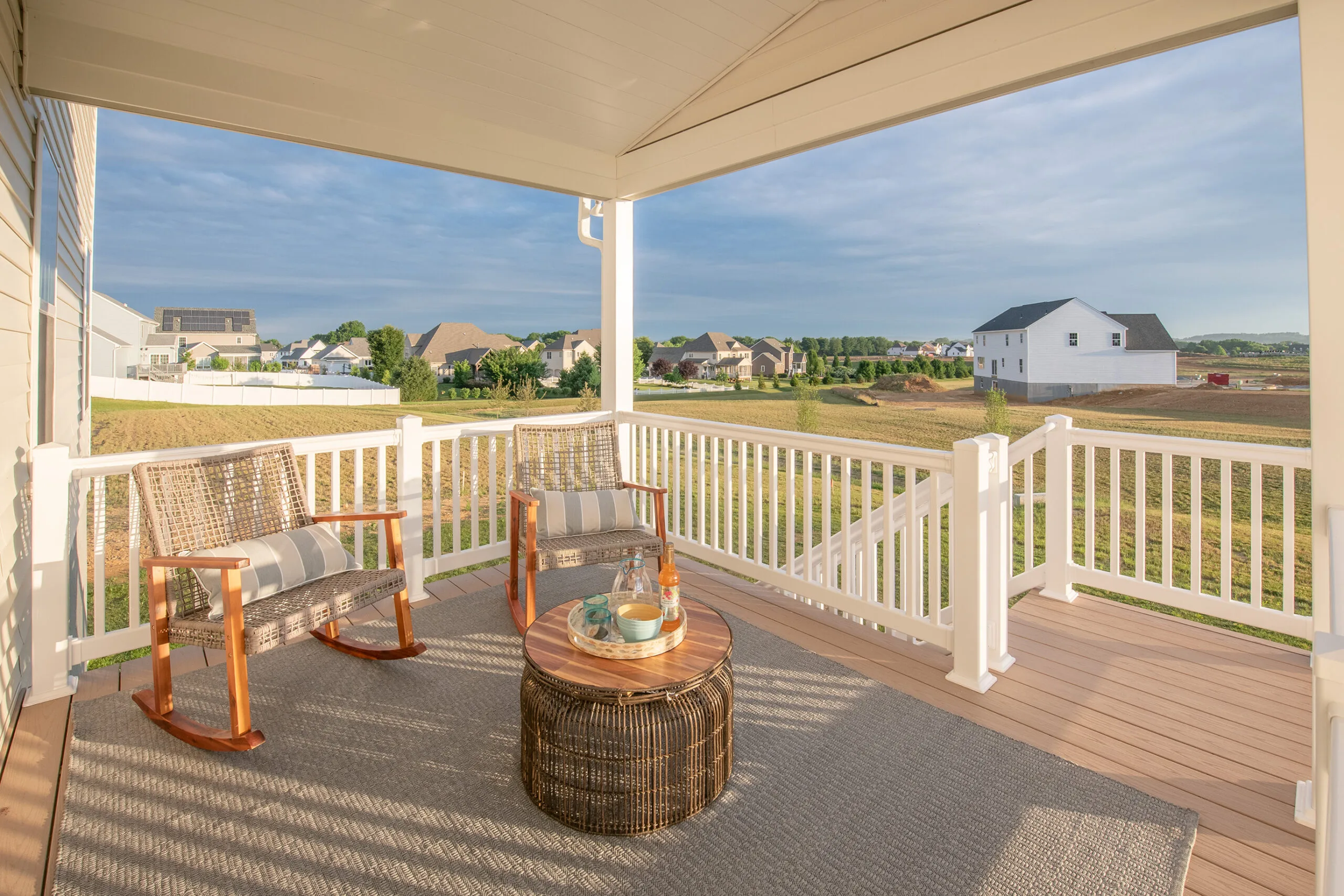 A covered porch with two wicker rocking chairs and a round wicker table on a rug, overlooking a grassy backyard and suburban houses under a partly cloudy sky.