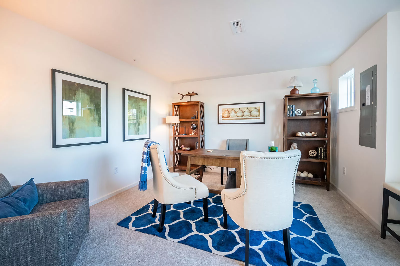 A bright home office with two white chairs, a wooden desk, a gray armchair, and wooden bookshelves on beige carpet. Blue and white rug, framed wall art, and decorative items create a cozy, modern feel.