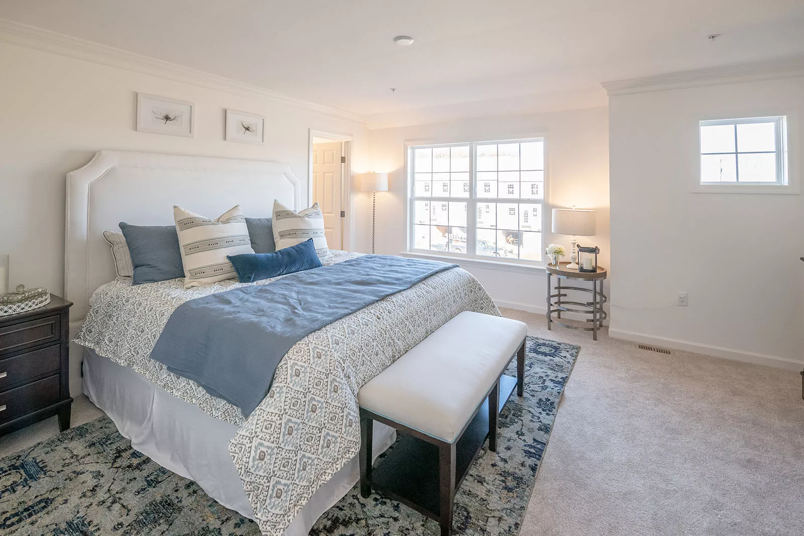 Bright bedroom with a large bed featuring patterned bedding and blue accents, a cushioned bench at the foot, dark bedside tables with lamps, large windows, and neutral walls with framed art above the bed.