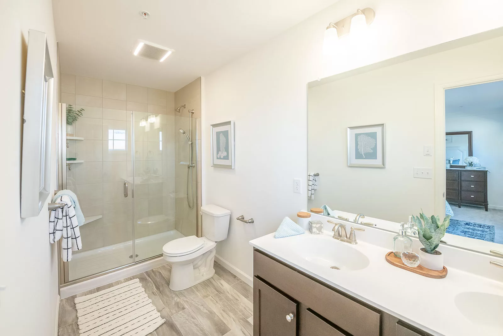 Bright, modern bathroom with a glass-enclosed shower, white toilet, double sink vanity with a large mirror, light wood floors, and minimal decor including towels, plants, and framed art on the walls.