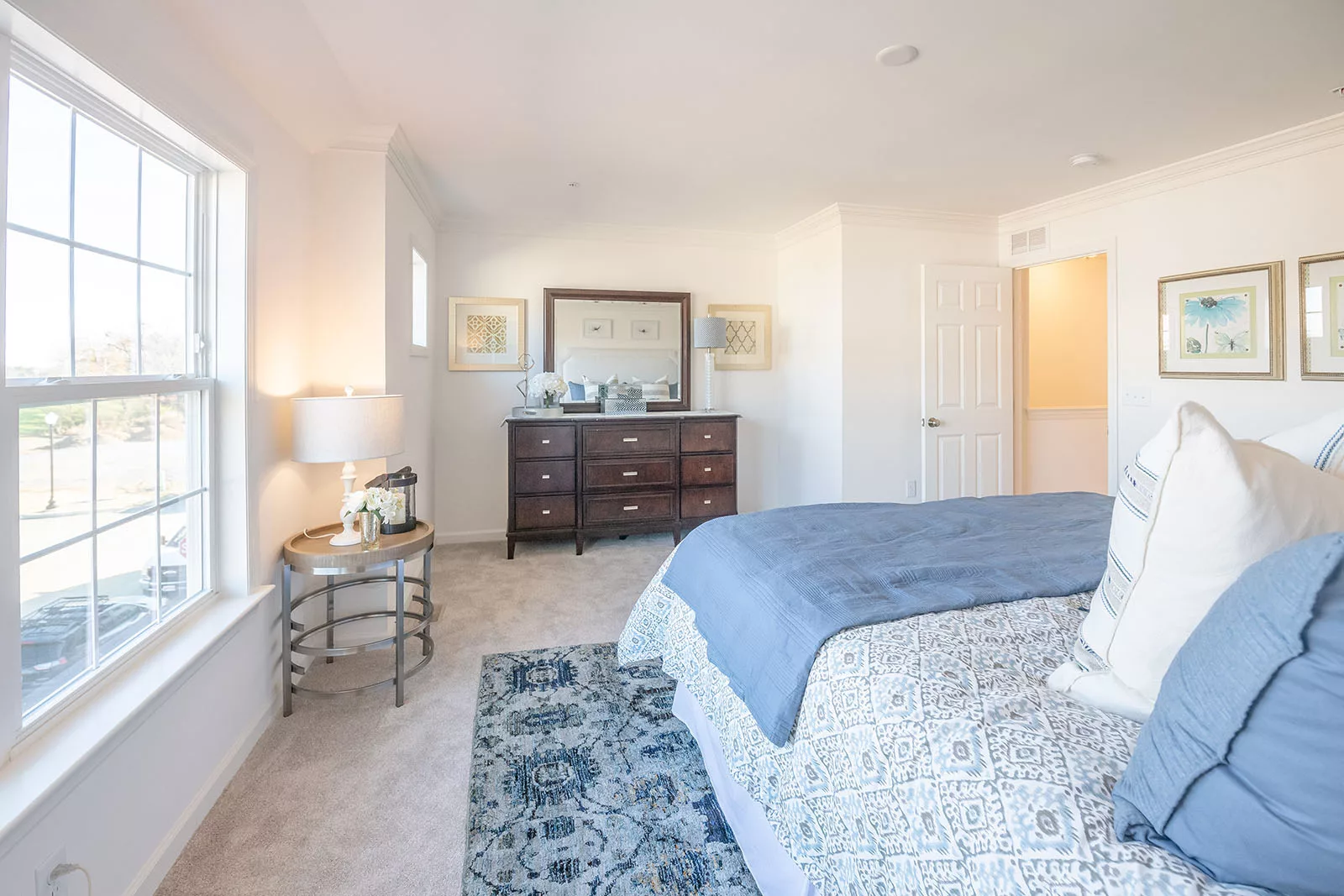 Bright bedroom with a large window, bed with blue and white bedding, patterned rug, dresser with mirror, table lamp, and framed artwork on white walls. The room appears clean and airy.