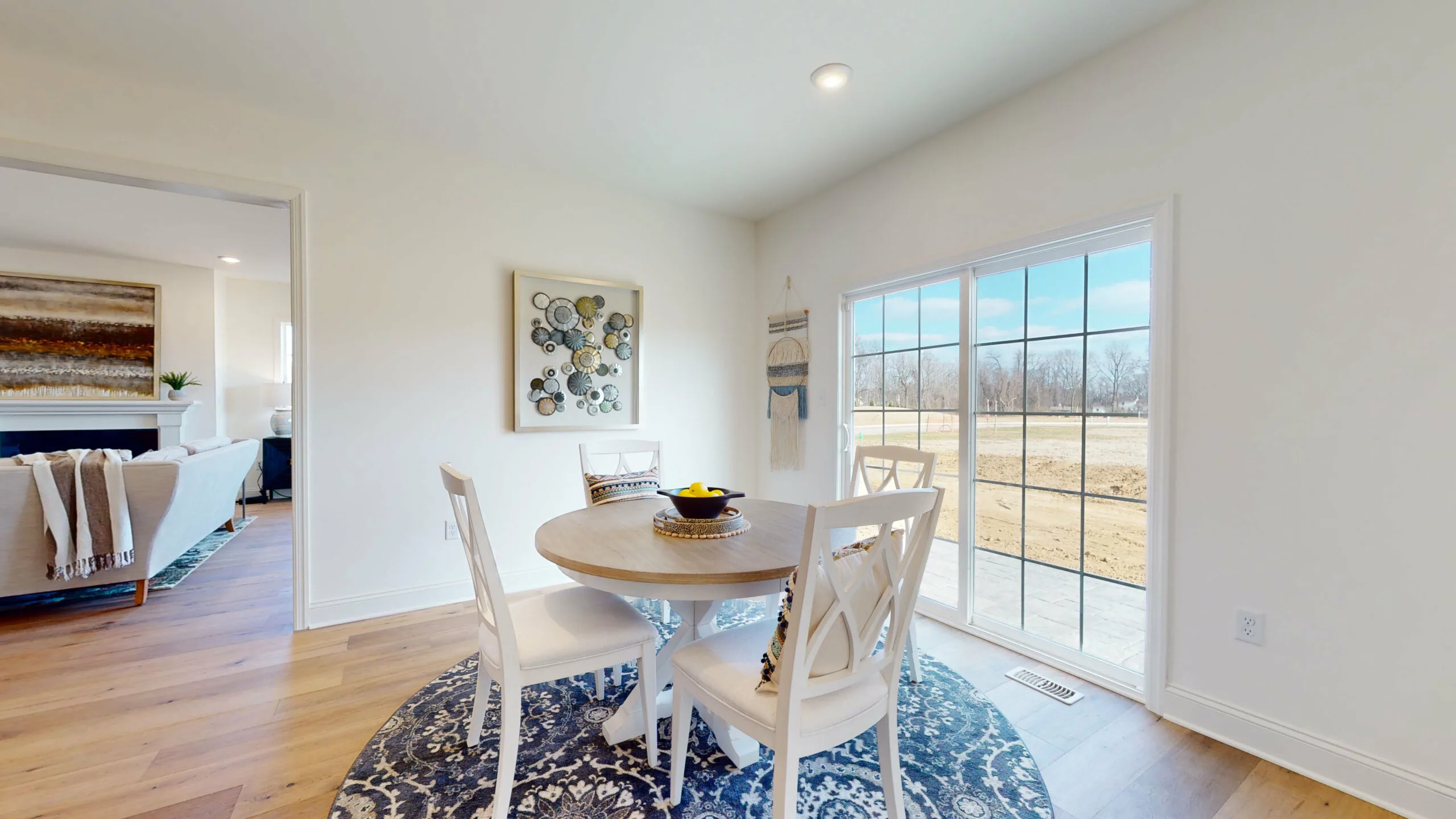 Bright dining area with a round table and four white chairs on a blue patterned rug, near large sliding glass doors overlooking a backyard. Art and decor adorn the white walls; a living room is visible through an open doorway.
