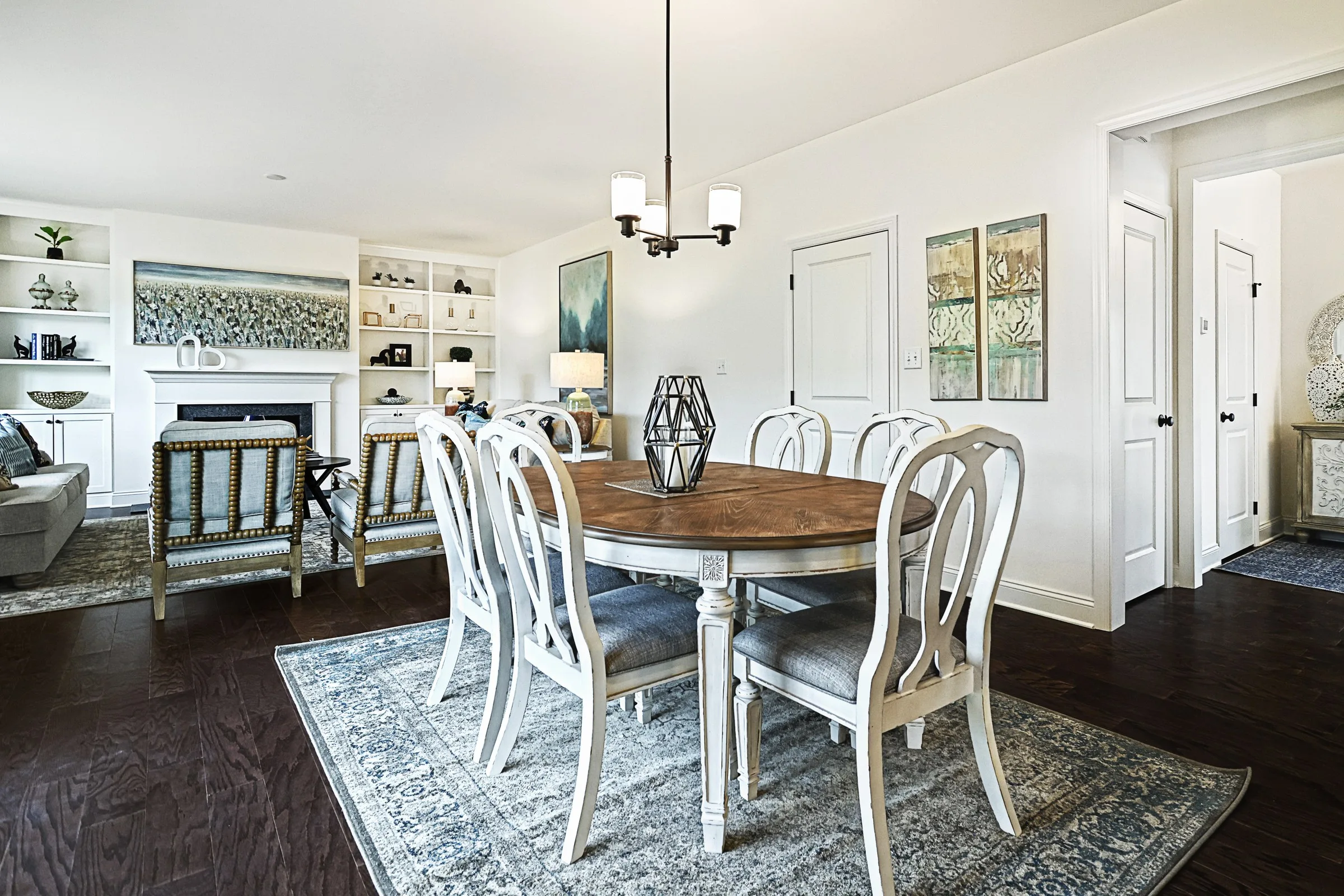 Bright dining room with a wooden table and six white chairs on a patterned rug. Open layout shows a living area with sofas, shelves, and wall art in the background. Modern light fixture hangs above the table.