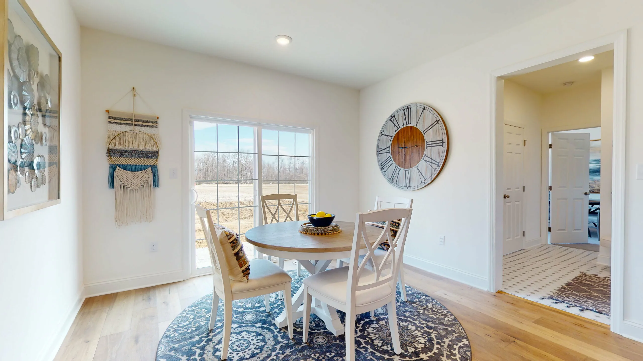 A bright dining area with a round table and four chairs sits on a patterned rug near sliding glass doors. A large wall clock, woven wall hanging, and framed art decorate the light, airy space.
