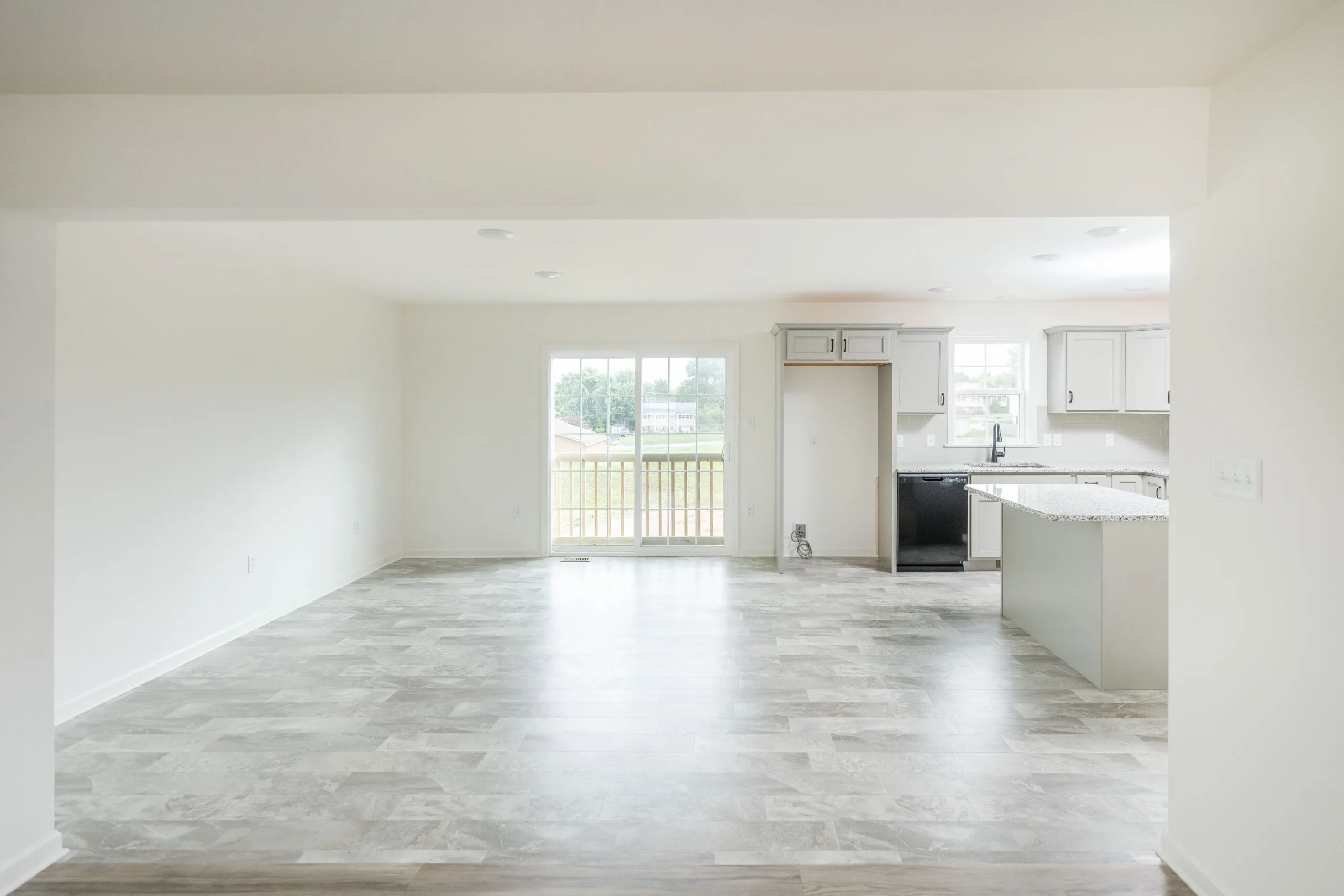 Bright, empty open-concept room with large windows, light-colored tile floors, white walls, and a modern kitchen area featuring an island, cabinets, and stainless steel appliances.