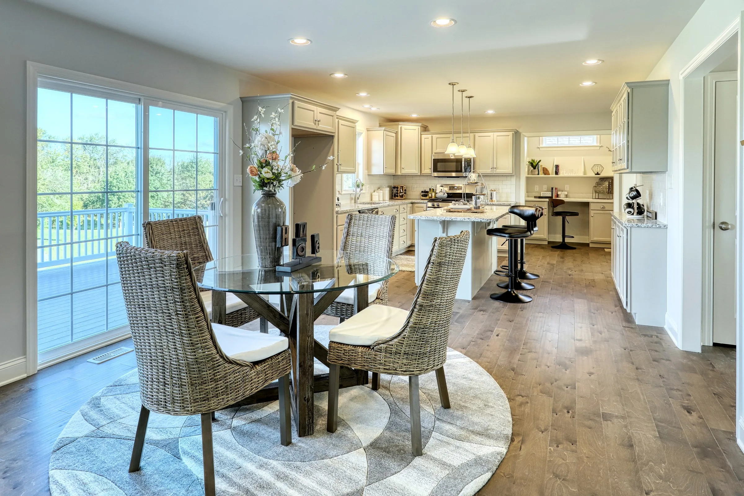 Modern kitchen and dining area with glass table, four wicker chairs on a round rug, pendant lights, bar stools at a kitchen island, white cabinetry, stainless steel appliances, and large sliding glass doors to a deck.
