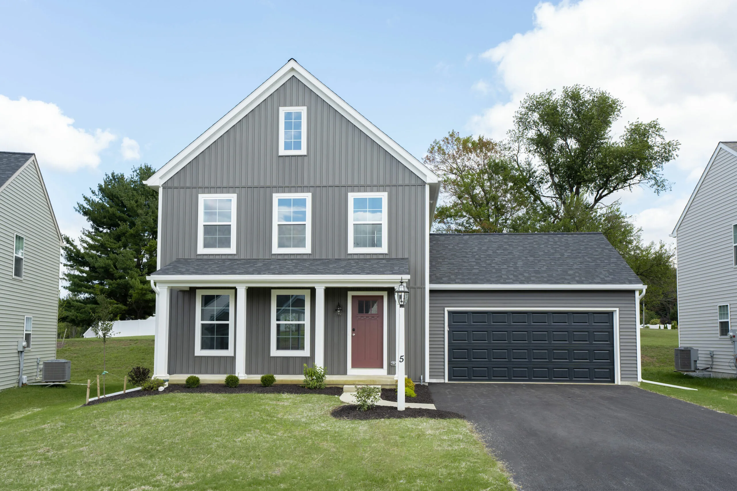 A modern two-story gray house with white trim, a covered front porch, a red front door, and an attached double garage, set on a green lawn with a driveway and trees in the background under a blue sky.