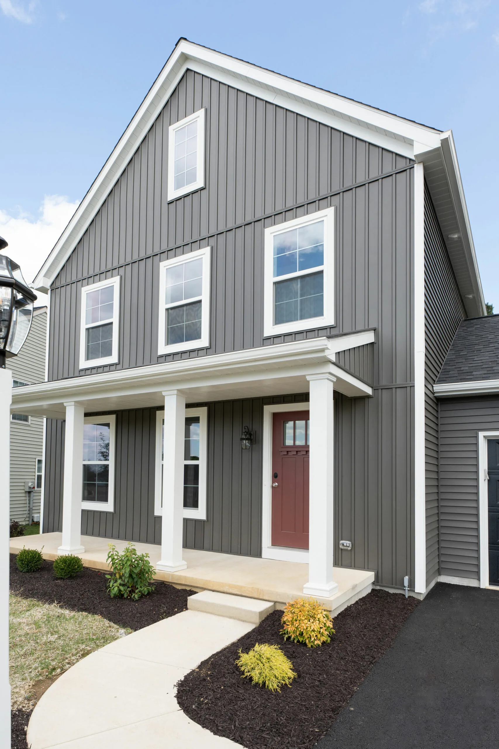 A modern two-story house with gray vertical siding, white trim, and a red front door. The home features a covered front porch with white columns and a landscaped yard with bushes and a curved walkway.