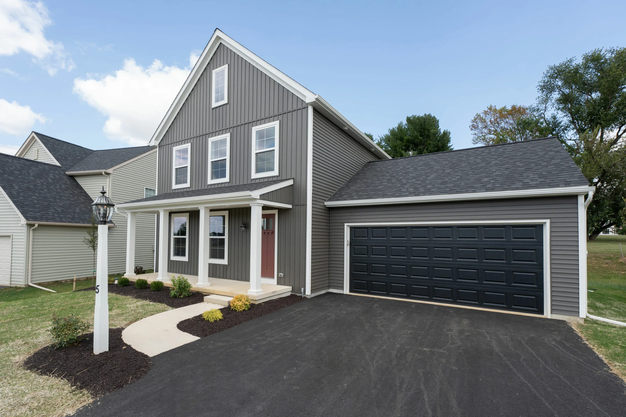 A modern two-story gray house with white trim, a covered front porch, and a large black garage door. The home has a paved driveway, small landscaped bushes, and a well-kept lawn under a blue sky.