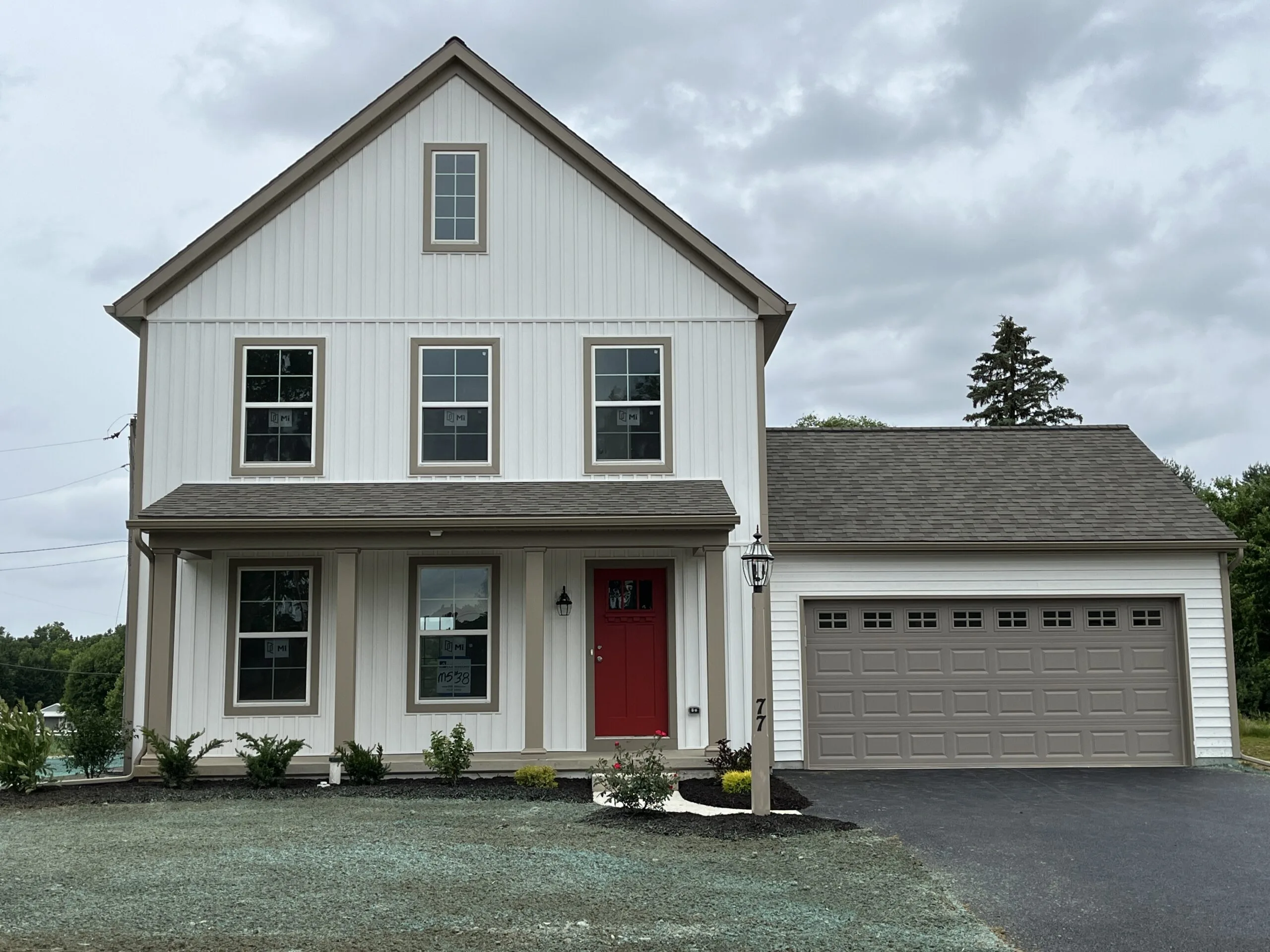 A modern two-story house with white siding, a red front door, and attached two-car garage. There are several windows, a small front porch, minimal landscaping, and a paved driveway under a cloudy sky.