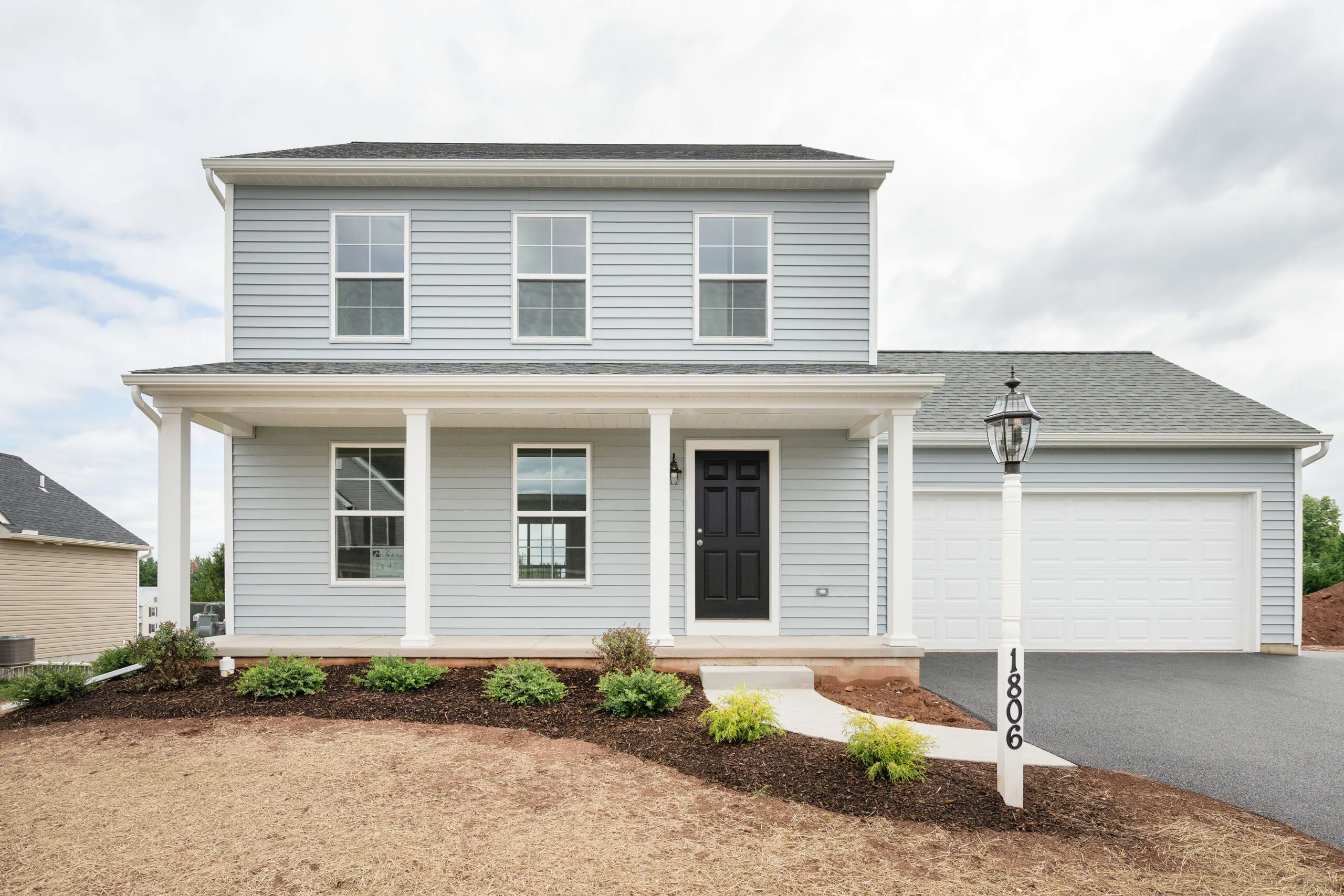 A two-story light blue house with white trim, a covered front porch supported by columns, a black front door, double garage, and a lamp post with the address number 1806. Small shrubs line the walkway.
