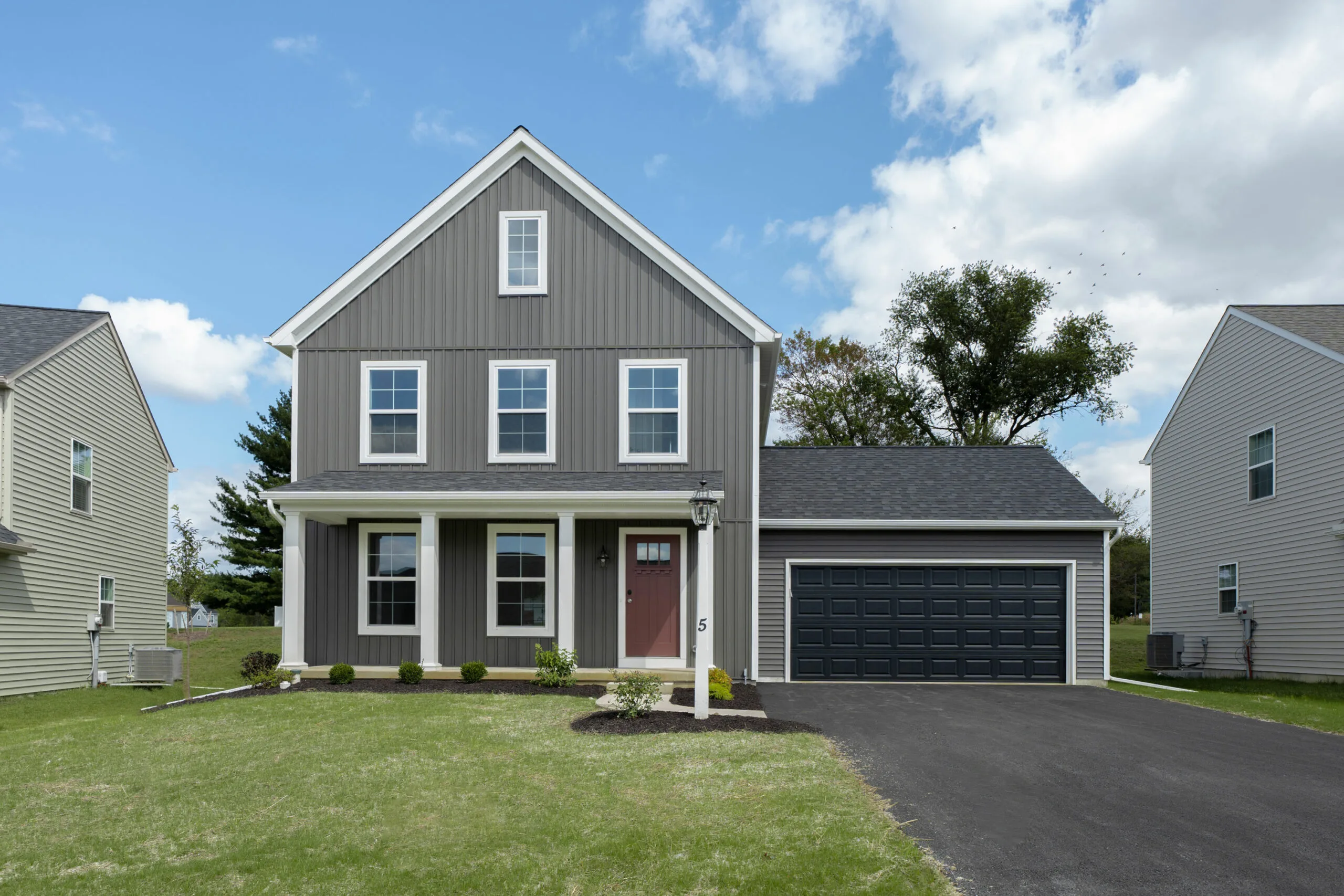 A modern two-story gray house with white trim, a front porch, and a dark garage door. The home has a manicured lawn, small shrubs, and a paved driveway, set against a blue sky with scattered clouds.