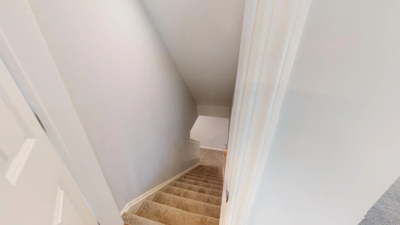 A view looking down a narrow, carpeted staircase with beige carpeting and white walls, seen from the top of the stairs near an open door.