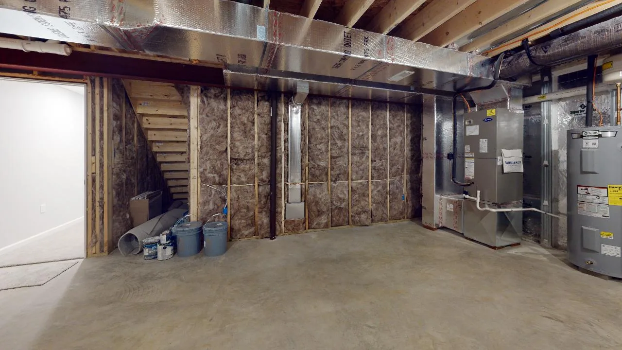 Unfinished basement with exposed insulation, pipes, and ductwork. Several paint cans and construction materials are on the floor near wooden stairs. A water heater and HVAC system are installed on the right side.