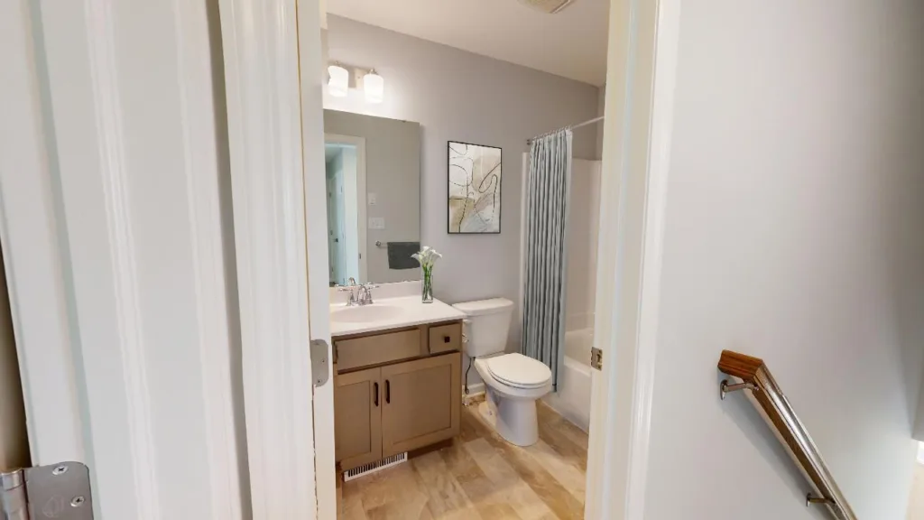 A modern bathroom with a light wood vanity, sink, mirror, and lights above. There is a white toilet, a shower with a patterned curtain, a vase with flowers on the counter, and artwork on the wall.