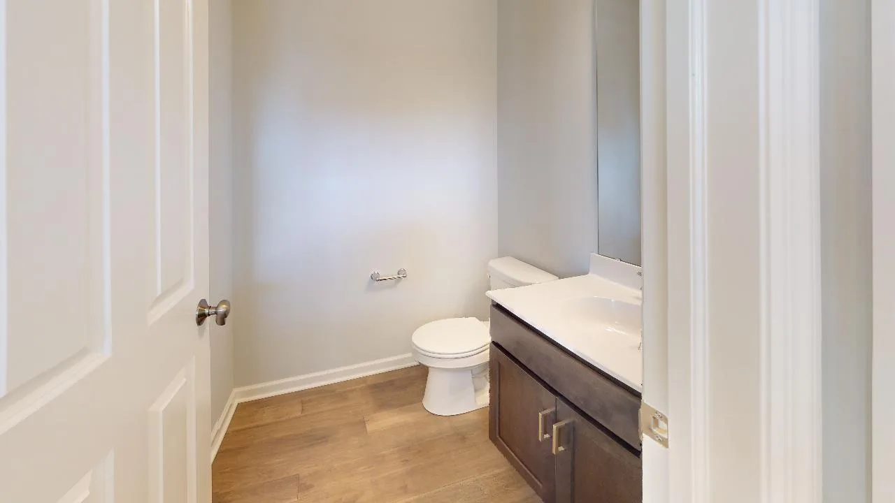 A small, simple bathroom with light-colored walls, a white toilet, a wall-mounted toilet paper holder, a dark wood vanity with a white countertop, and a large mirror above the sink. The floor is light wood.