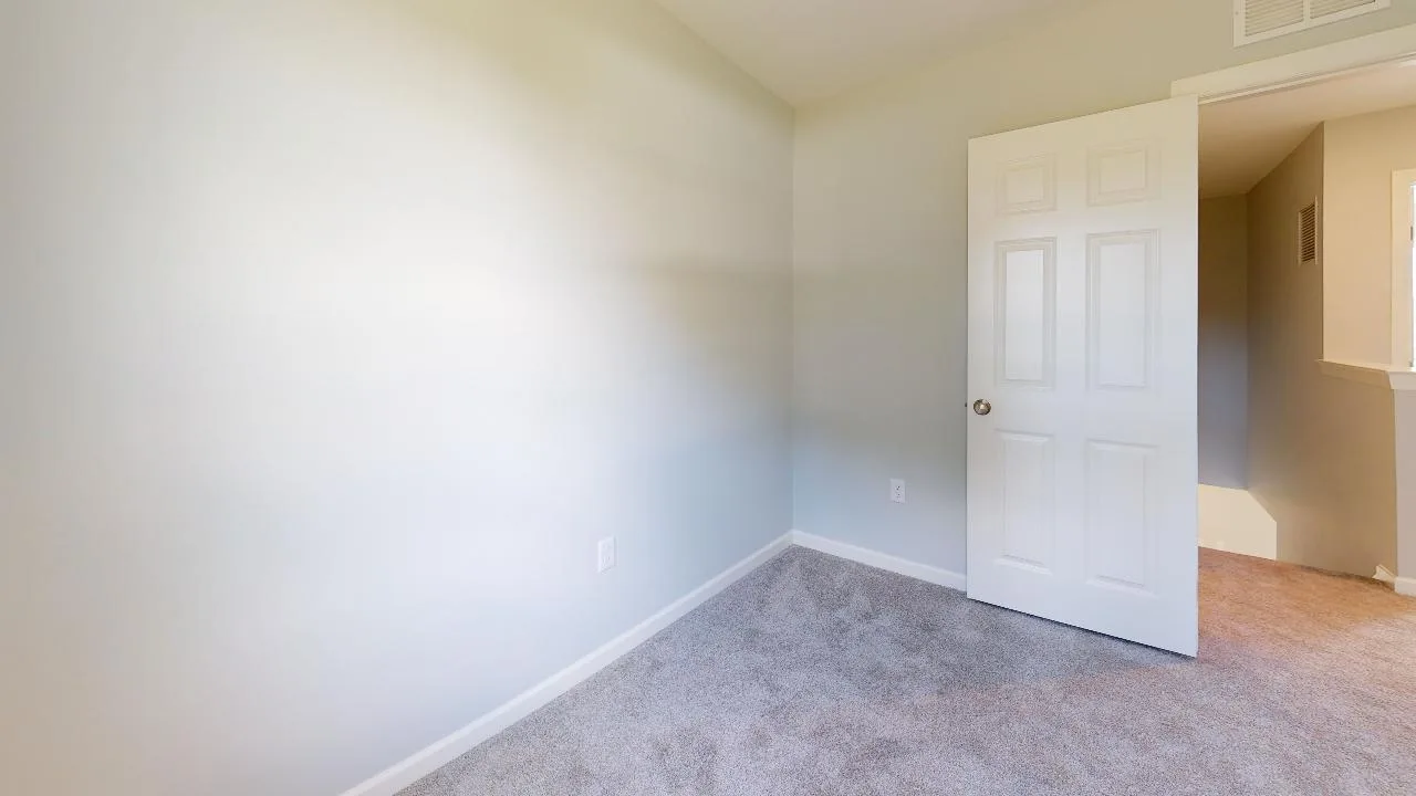 A small empty room with beige carpet, light gray walls, and an open white door leading to a hallway with similar carpeting. The room is well-lit by natural light.
