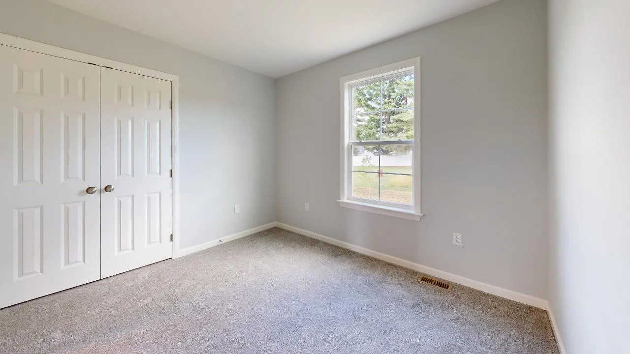 A small empty room with light gray walls, a beige carpet, a double-door closet, and a single window showing trees and grass outside. Natural light brightens the space.