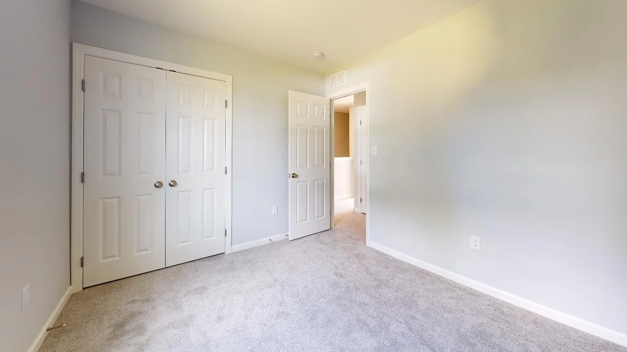 Empty bedroom with light gray carpet, white walls, two closed double-door closets on the left, and an open door leading to a hallway with natural light.