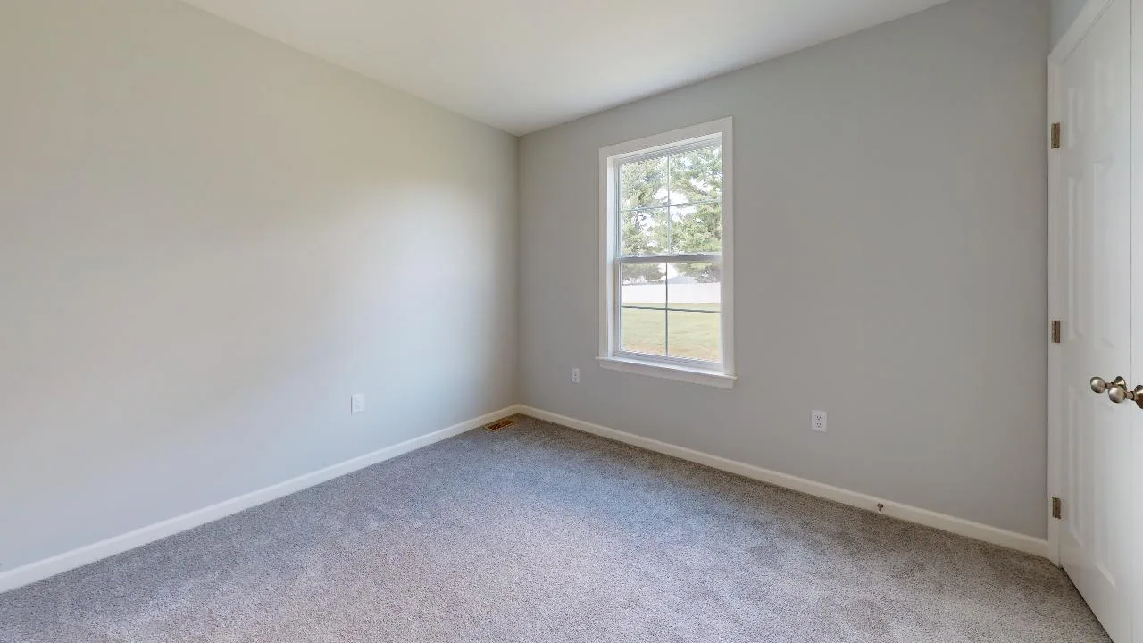 An empty room with light gray walls, a single window letting in natural light, carpeted flooring, and a white door with a round handle.