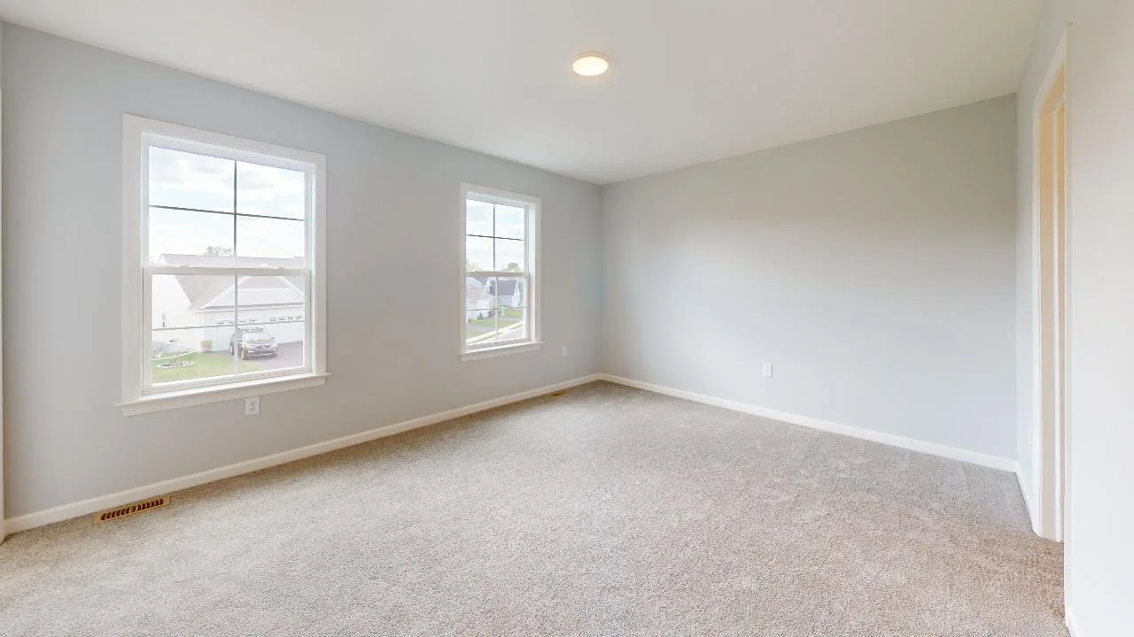 A bright, empty room with two large windows, light gray walls, beige carpet flooring, and a ceiling light. Sunlight enters through the windows, illuminating the clean, spacious area.