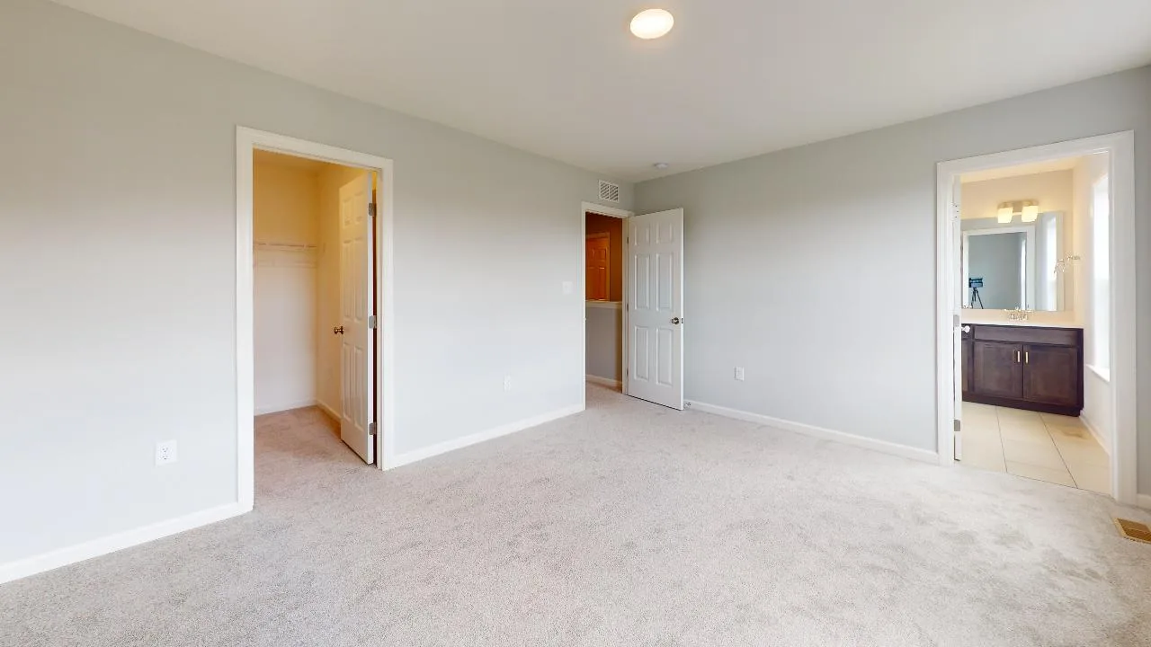 A bright, empty bedroom with light gray walls, beige carpet, and three doors—one to a walk-in closet, one to a hallway, and one leading to a bathroom with a vanity and mirror visible.