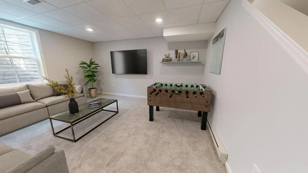 A modern basement recreation room with beige carpet, a sectional sofa, glass coffee table, wall-mounted TV, foosball table, potted plant, and wall art under a drop ceiling with recessed lighting.