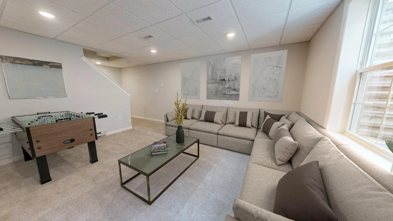 A cozy basement room with a large beige sectional sofa, a glass coffee table with a flower vase, foosball table, abstract wall art, carpeted floor, and a window letting in natural light.