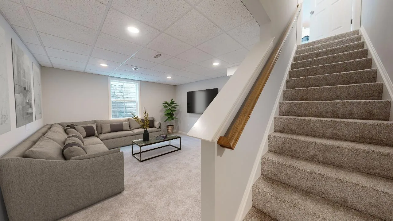 A modern basement living room with a large gray sectional sofa, glass coffee table, wall-mounted TV, potted plant, light carpet, and a staircase leading up. Artwork decorates the light-colored walls.
